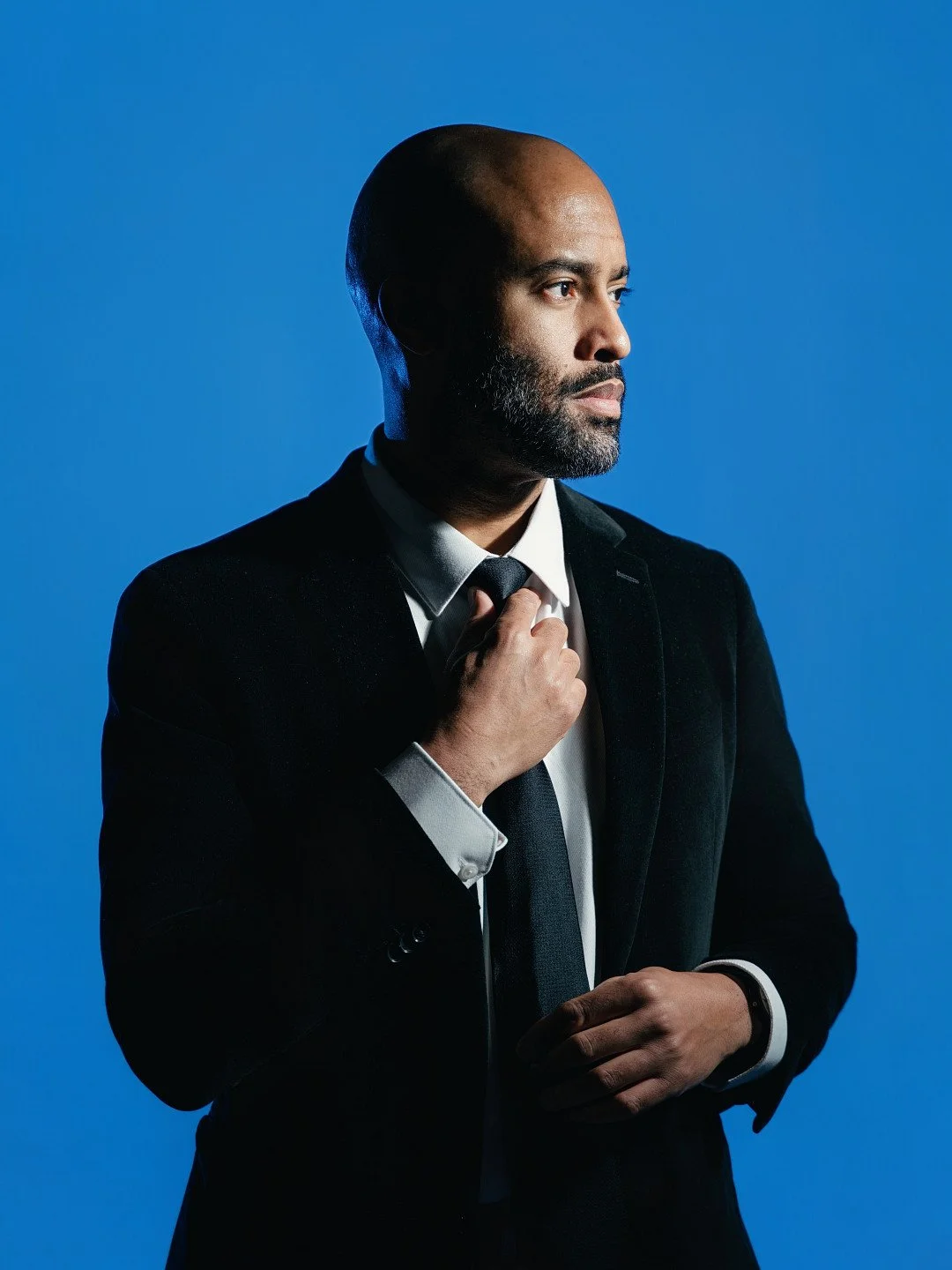 A man in a black suit and tie adjusting his tie against a blue background.