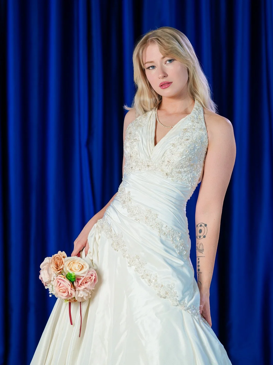 A young woman in a white wedding dress holding a bouquet of pink and peach roses stands in front of a blue curtain.