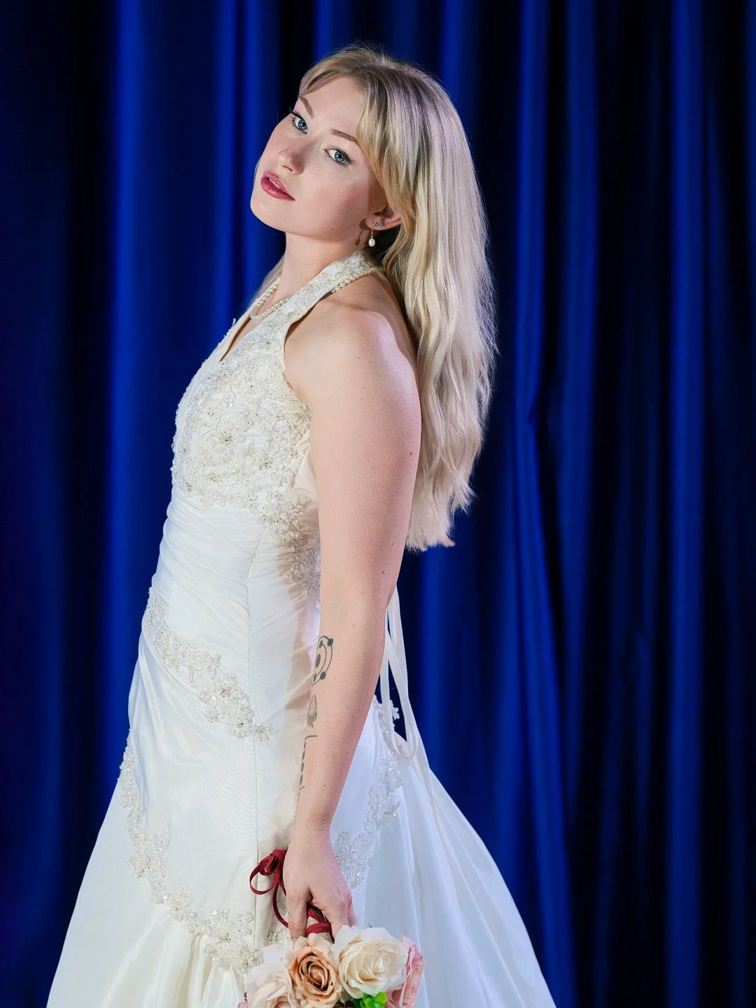 A woman in a white wedding dress holding a bouquet of roses standing in front of a blue curtain.