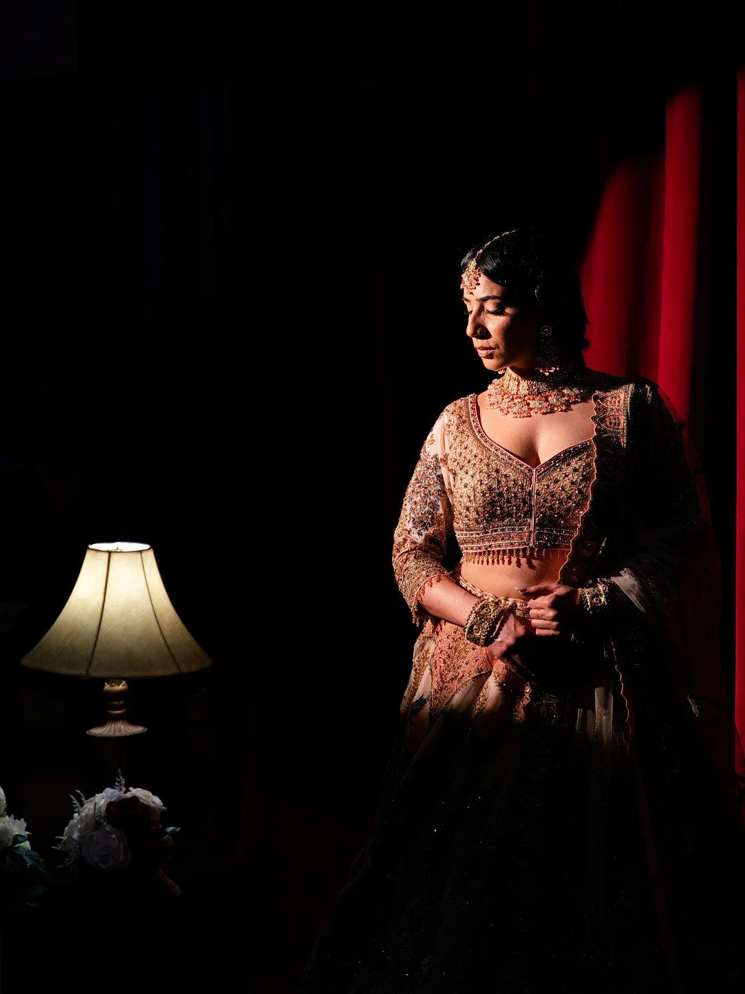 Woman dressed in traditional Indian wedding attire with jewelry, standing in low light near a lamp and flowers.
