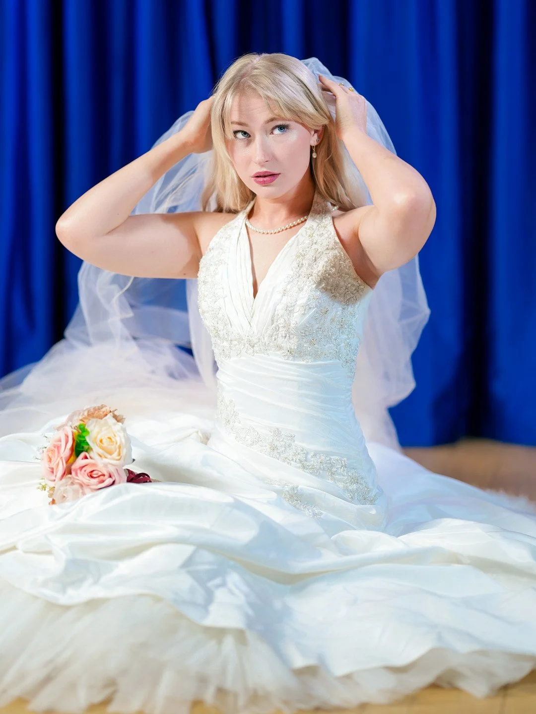 A woman in a wedding dress adjusting her veil, sitting in front of a blue curtain, with a bouquet of pink and cream roses on her lap.