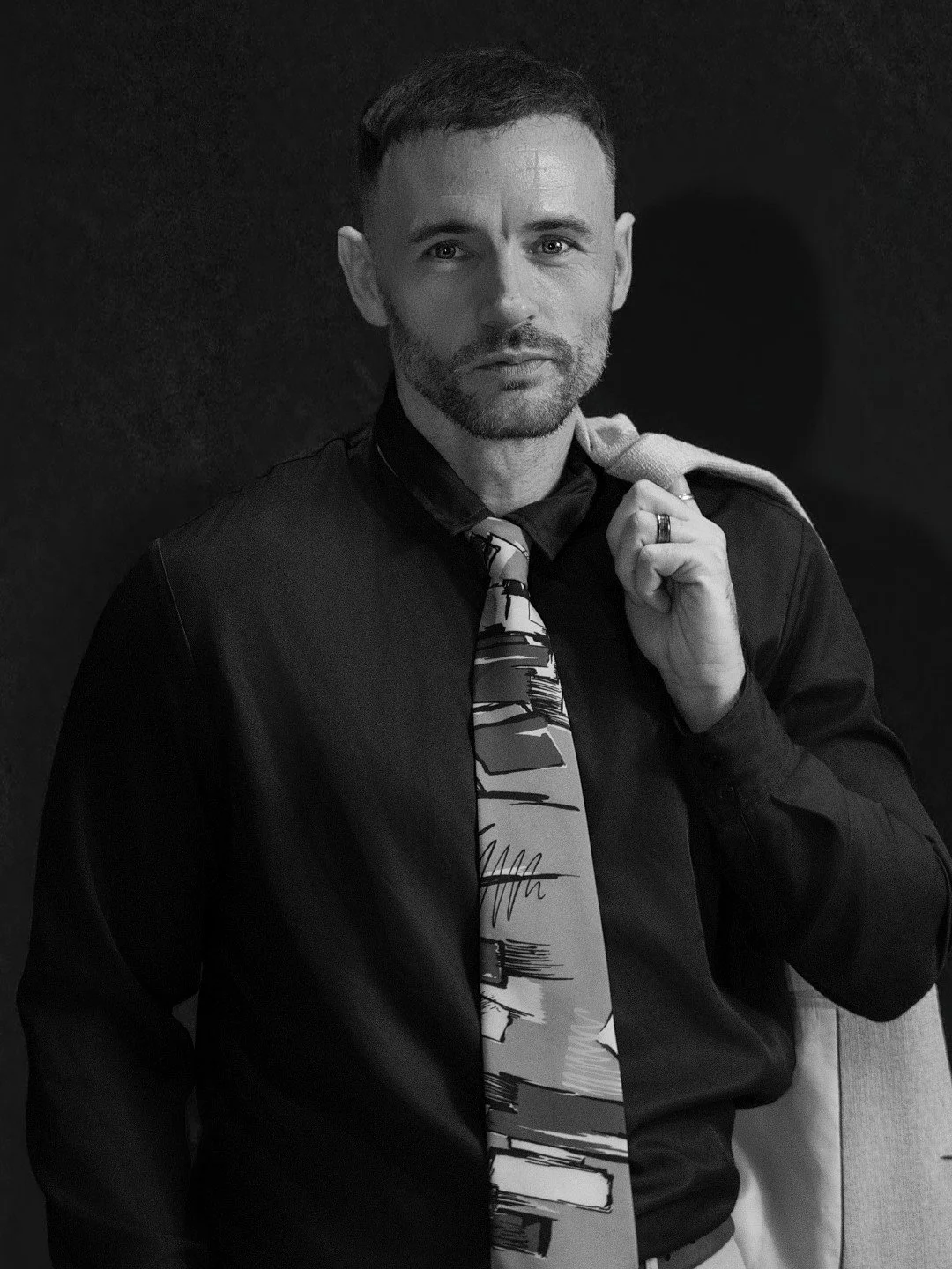 Black and white portrait of a man with short hair and a beard, wearing a dark shirt and patterned tie, holding a jacket over his shoulder.