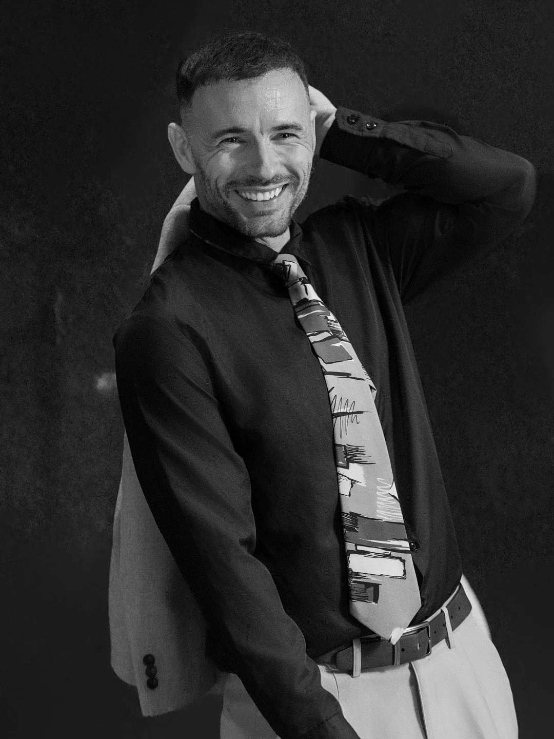 A smiling man with short hair, wearing a dark blazer, patterned tie, light-colored pants, and carrying a light-colored jacket over his shoulder, standing against a dark background.