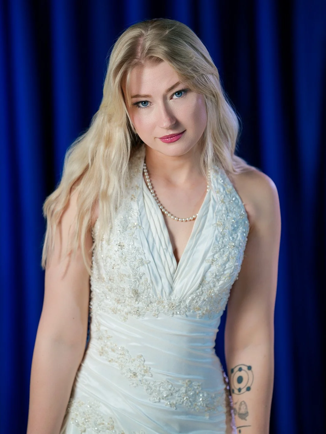 A young woman with long blonde hair, blue eyes, and fair skin wearing a white dress with lace details and pearls, posing in front of a blue curtain.