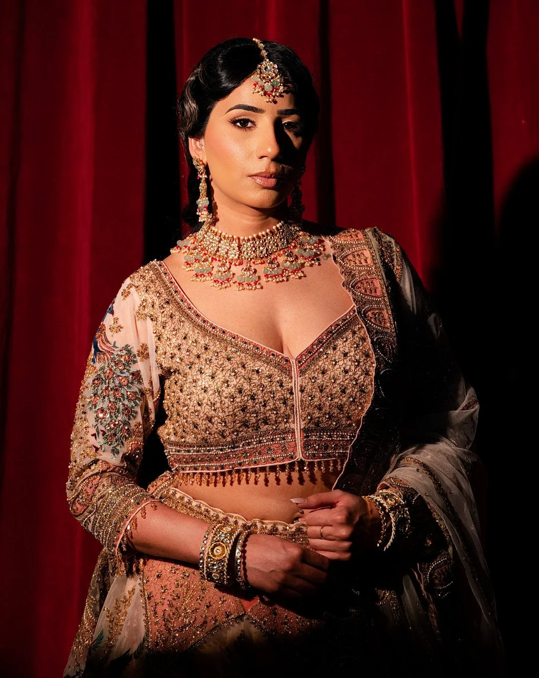 Woman dressed in a traditional Indian bridal outfit with intricate embroidery and jewelry, standing in front of a red curtain.