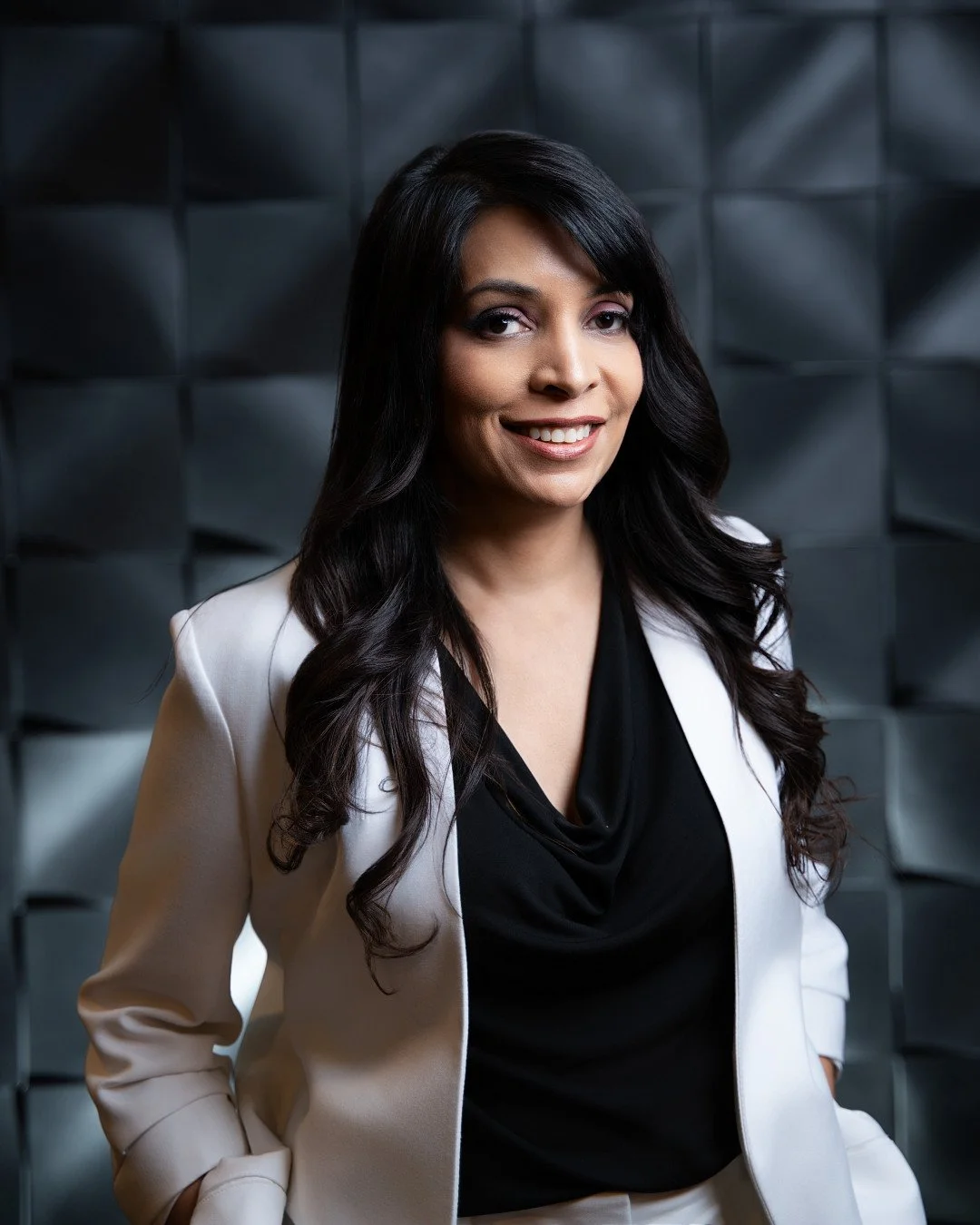Professional woman with long dark hair smiling, wearing a white blazer over a black top, standing in front of a textured gray wall.