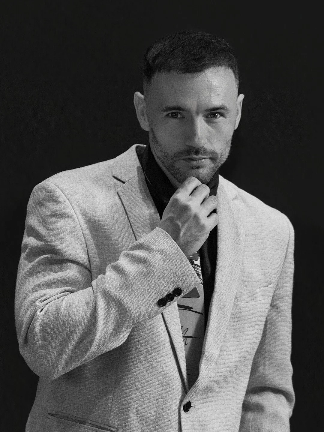 A black and white portrait of a man with short dark hair and facial hair, wearing a light-colored suit jacket over a dark shirt, looking directly at the camera with a hand touching his chin.