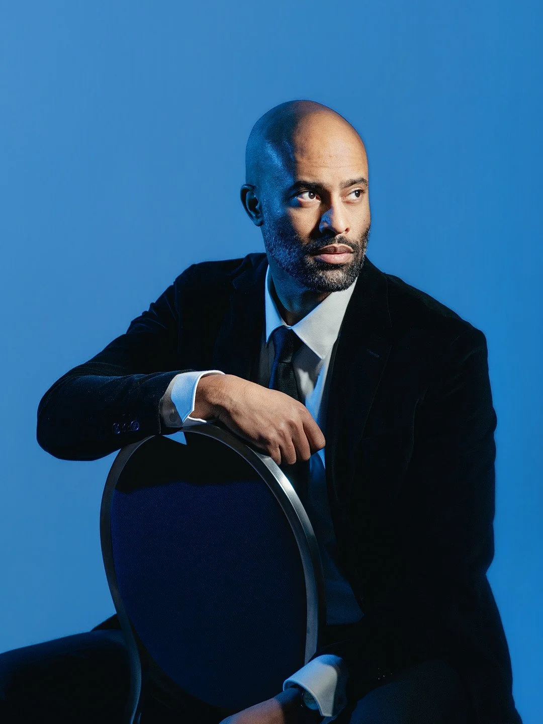 A man in a black suit and white shirt with a black tie, sitting on a chair against a blue background, looking to the side with a serious expression.