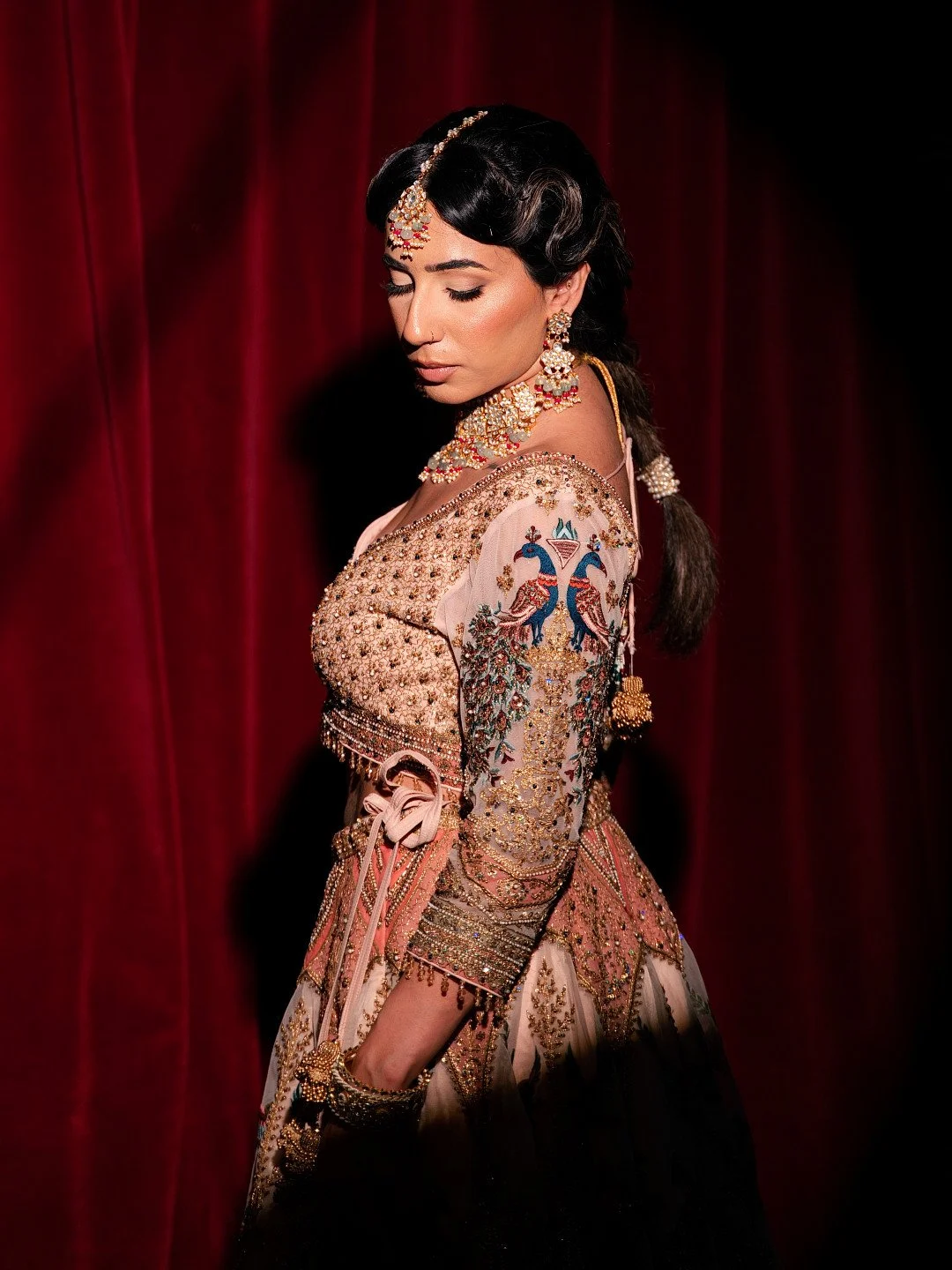 A woman wearing traditional, ornate Indian attire with detailed embroidery, jewelry, and a dark hairstyle, posing indoors against a red curtain.