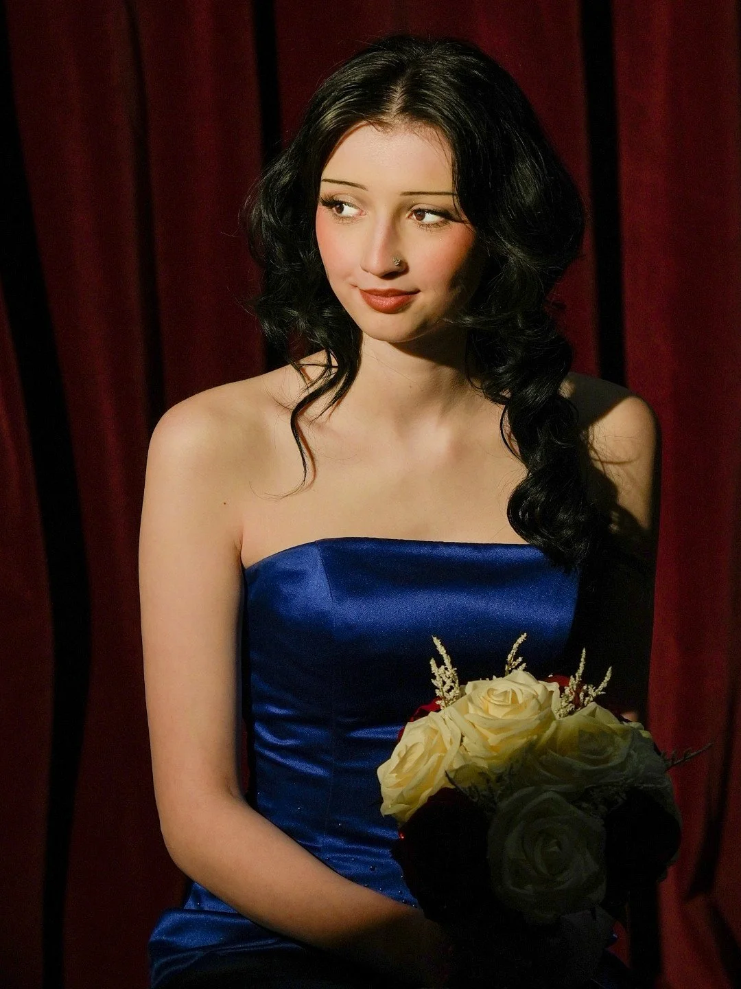 Young woman with black hair wearing a blue strapless dress, holding a bouquet of cream and black roses with other flowers, standing in front of red curtains.