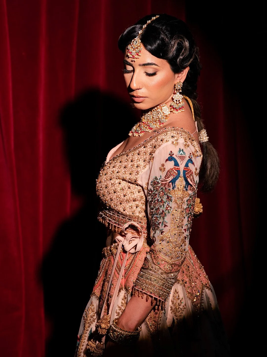 Woman wearing traditional Indian bridal attire with intricate embroidery, jewelry, and a red curtain background.