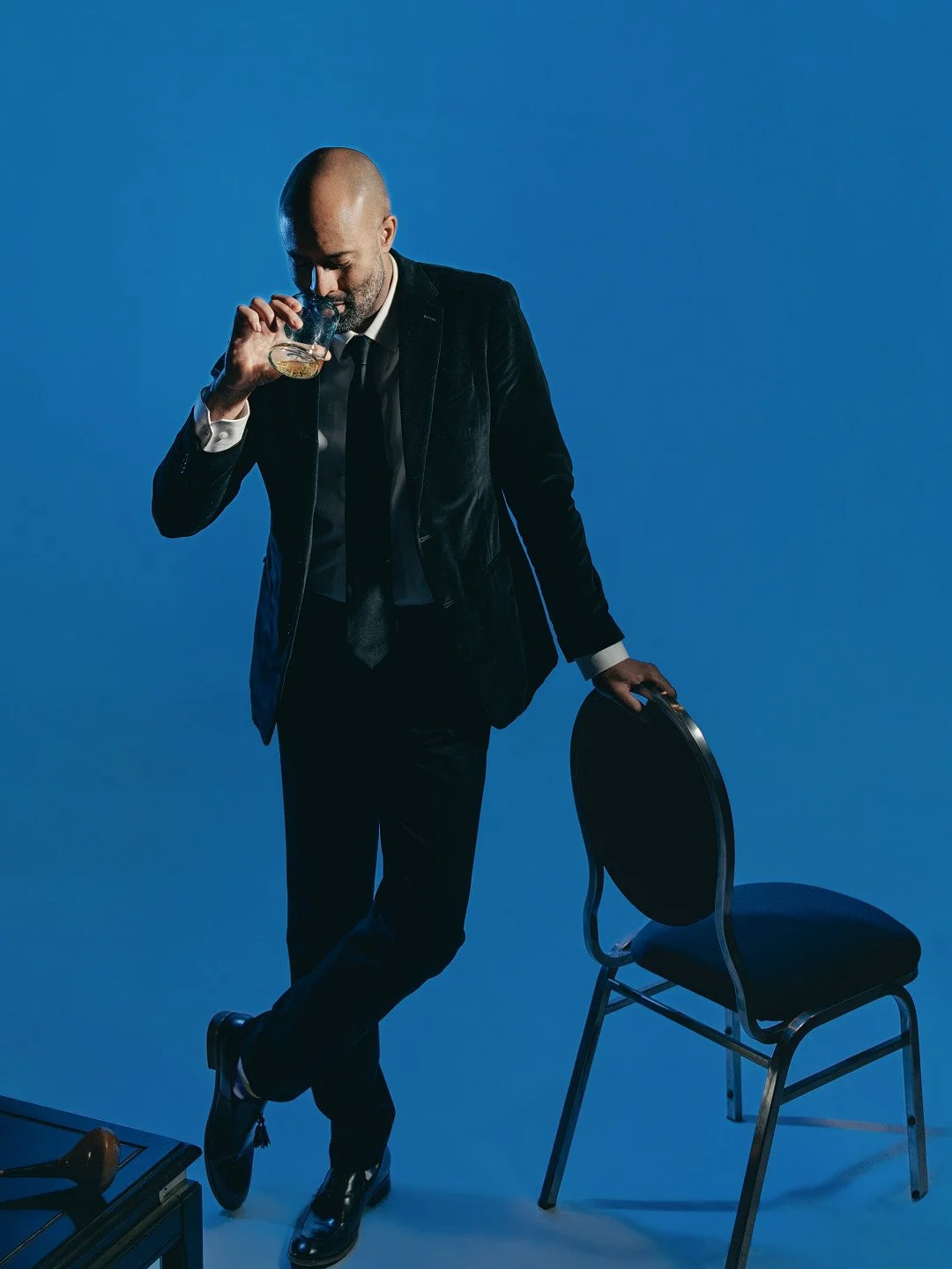 A man in a black suit and tie drinks whiskey while standing next to a chair, with a blue background.