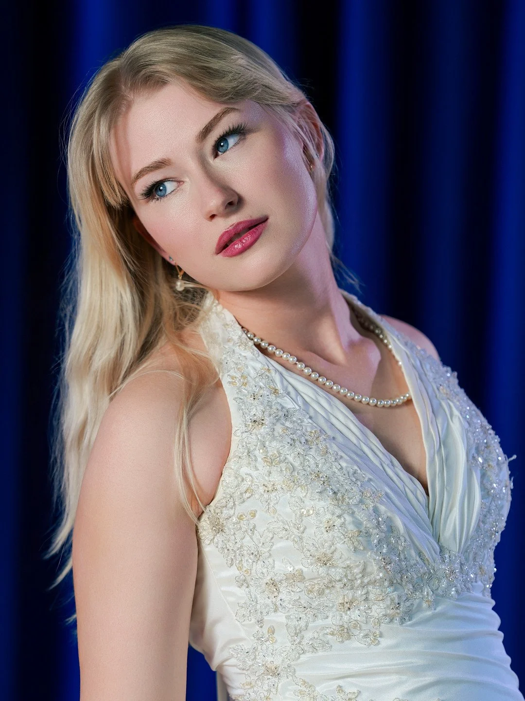 A woman with blonde hair and blue eyes, wearing a white dress with embroidery, pearl necklace, and earrings, standing against a blue curtain background.