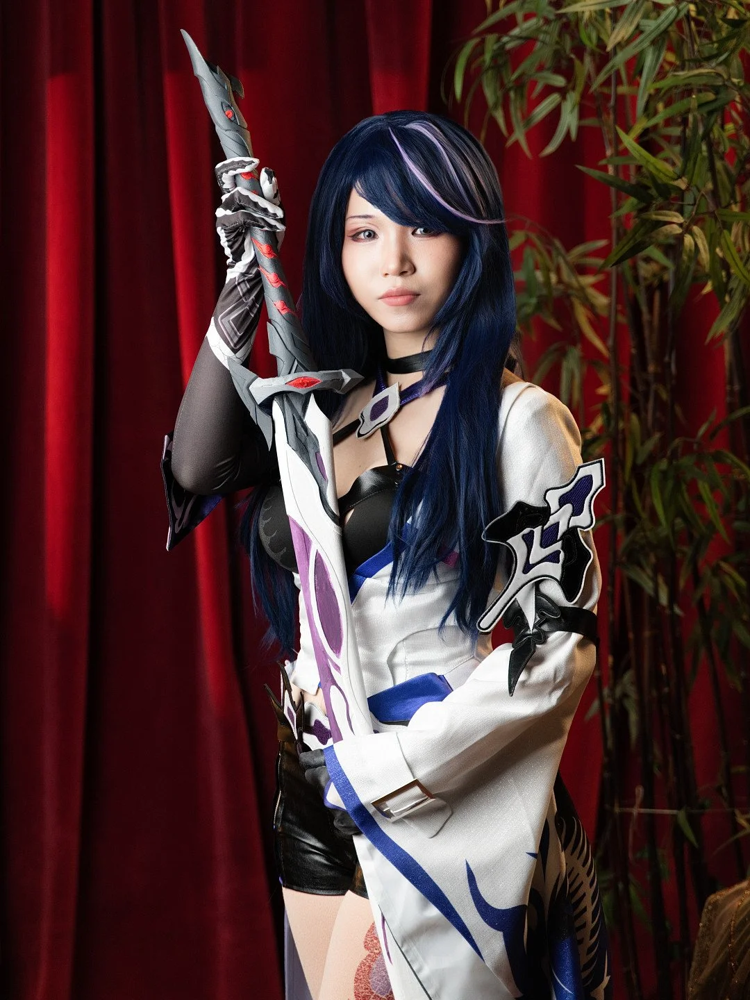 Cosplayer dressed as a character, holding a sword, with long dark blue hair with purple streaks, standing in front of red curtains and a potted plant.