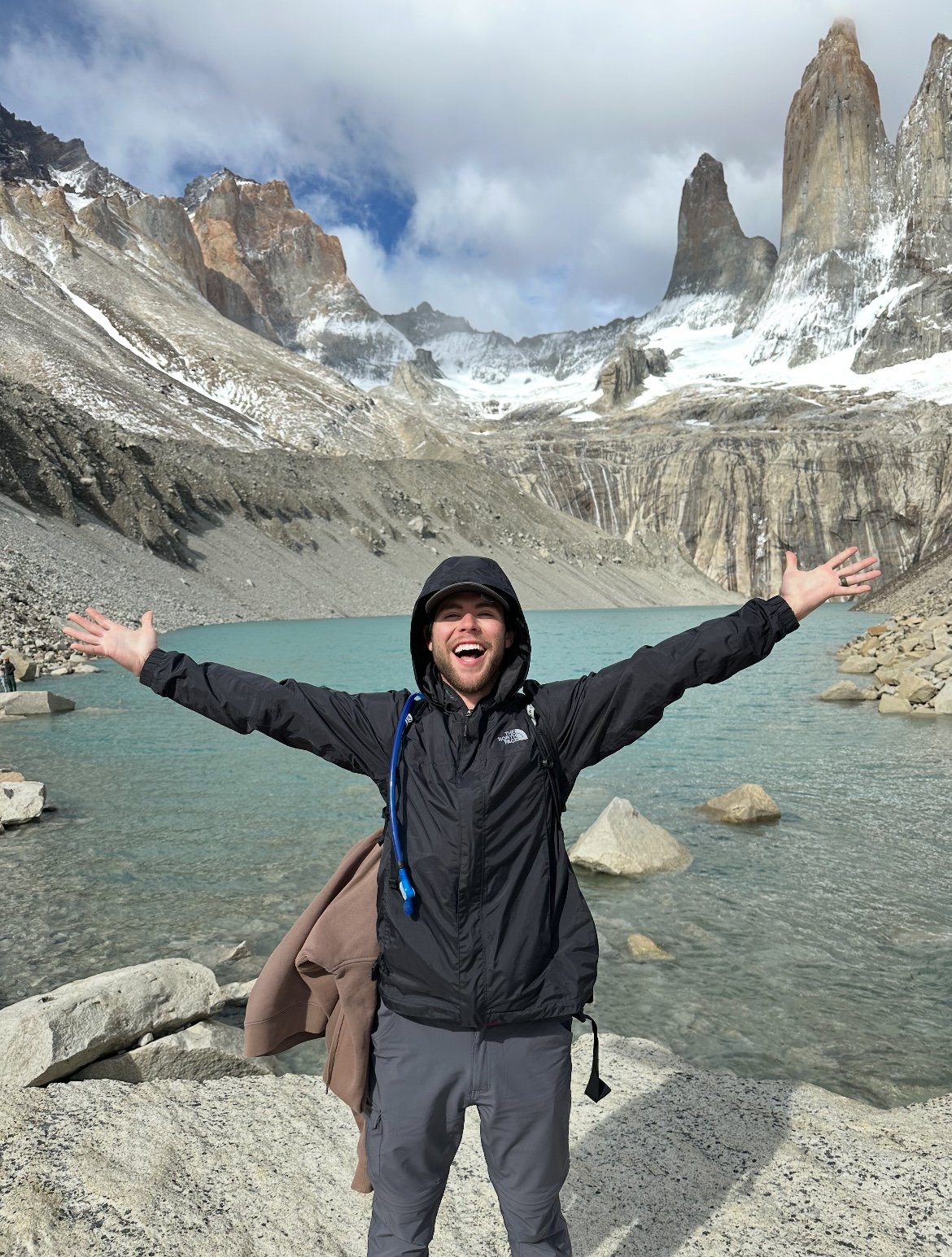 A man in outdoor gear with arms outstretched standing by a turquoise lake, surrounded by rocky mountains with snow and clouds in the background.