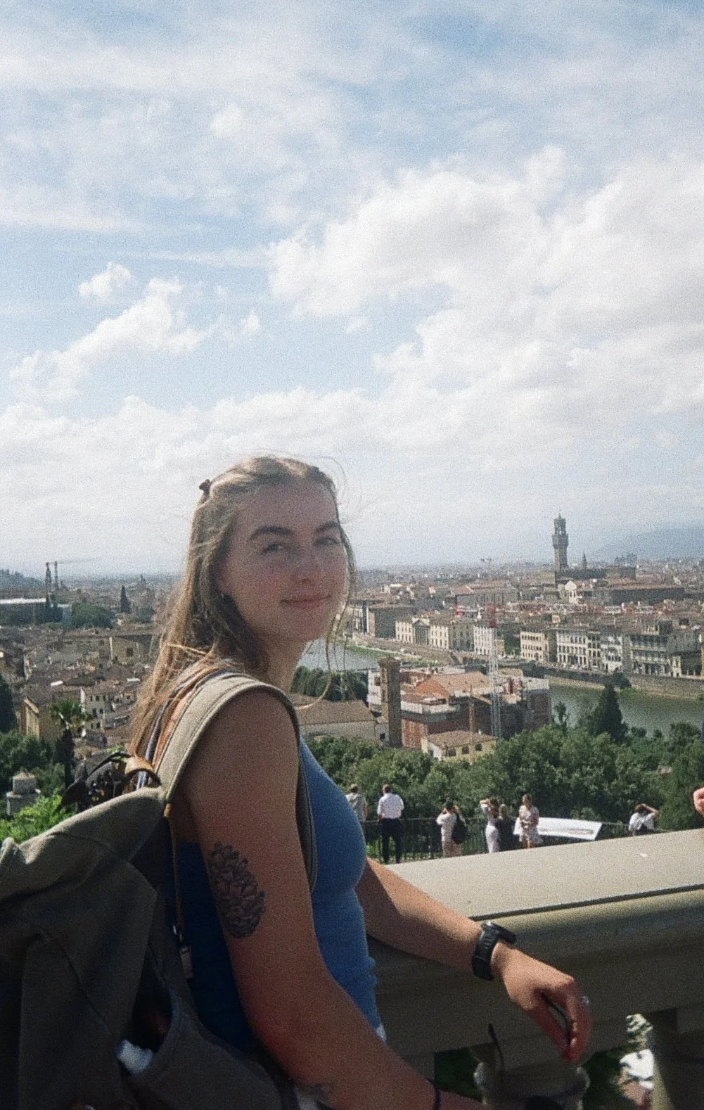 A young woman standing on a balcony with a scenic view of a historic European city, possibly Florence, Italy, with a river and a tall tower in the background on a partly cloudy day.