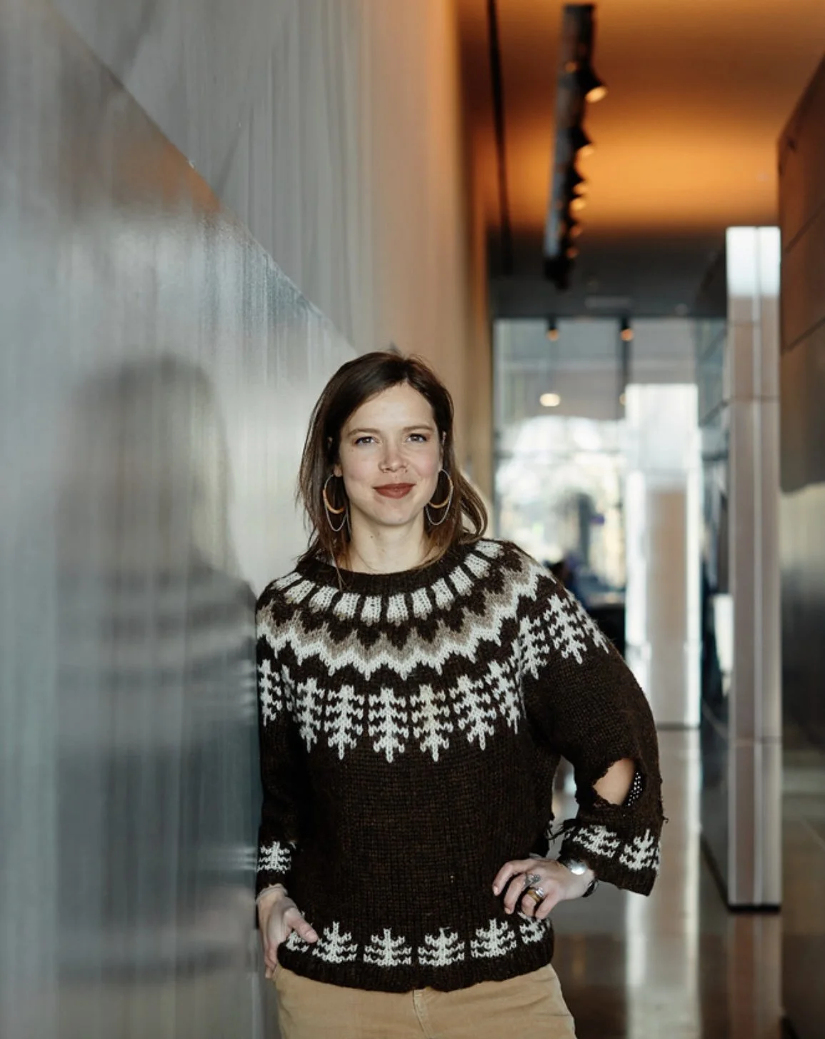 A woman standing in a modern indoor space, leaning against a wall, wearing a black and white patterned sweater and beige pants.