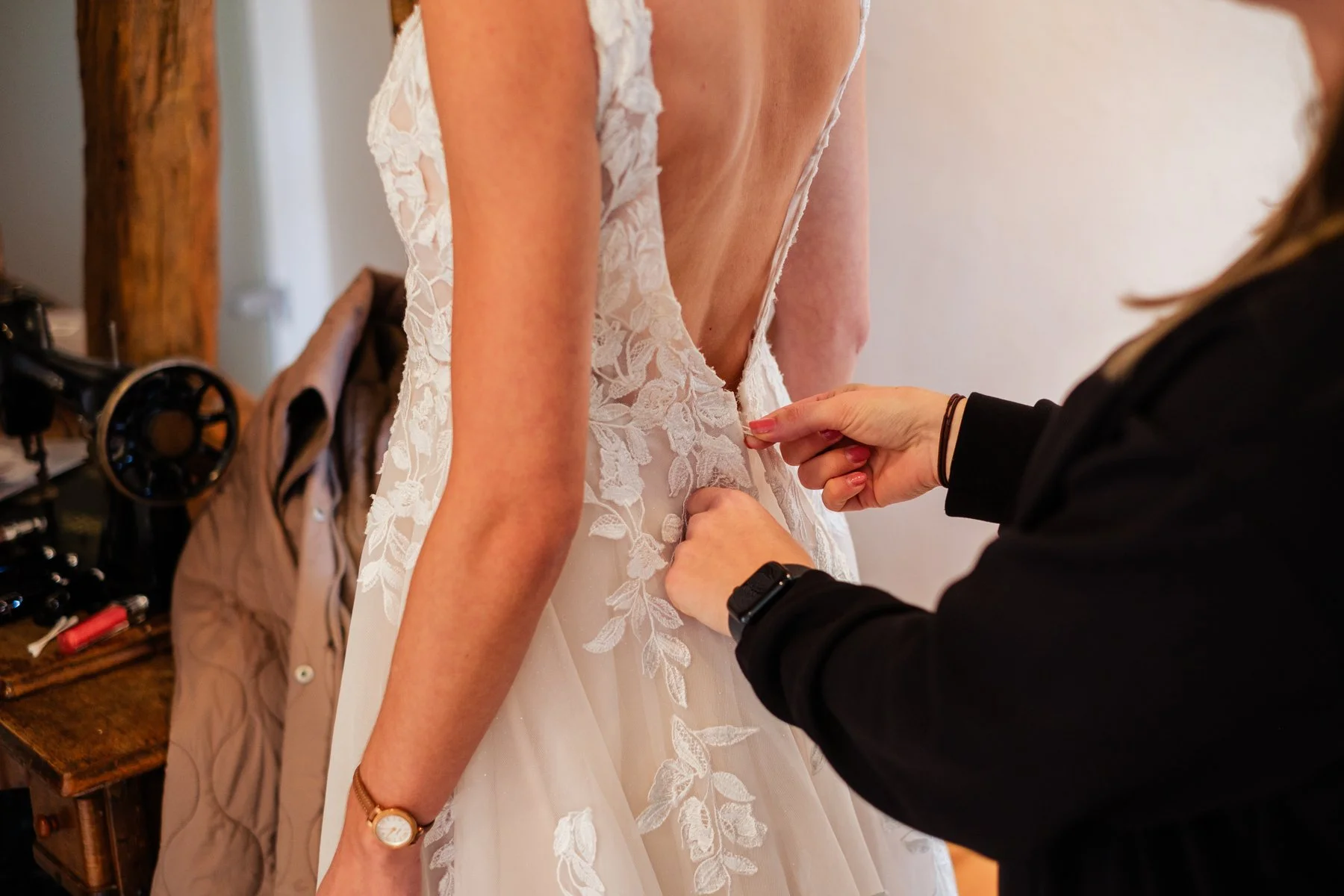 Eine Braut wird in einem weißen Hochzeitskleid mit Spitzen-Details von einer Person für die Zeremonie vorbereitet.