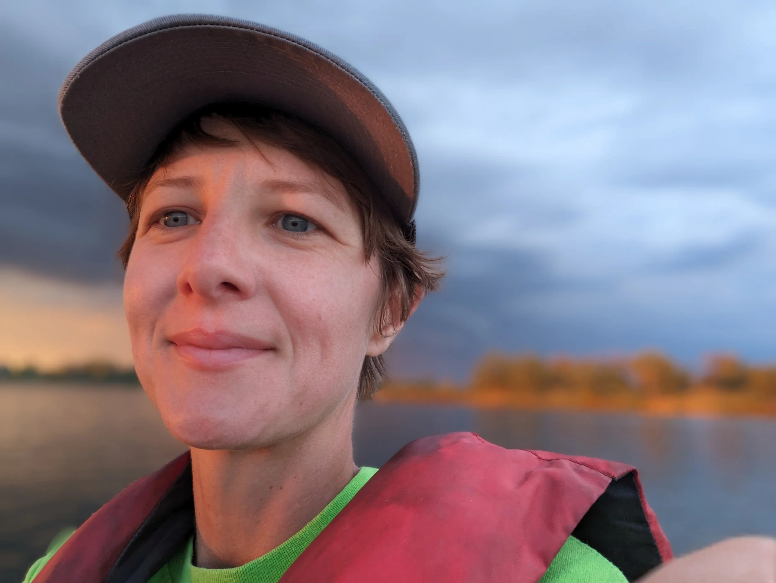 A young man with light skin, blue eyes, and brown hair wearing a cap and a life jacket on a boat during sunset with a cloudy sky in the background.