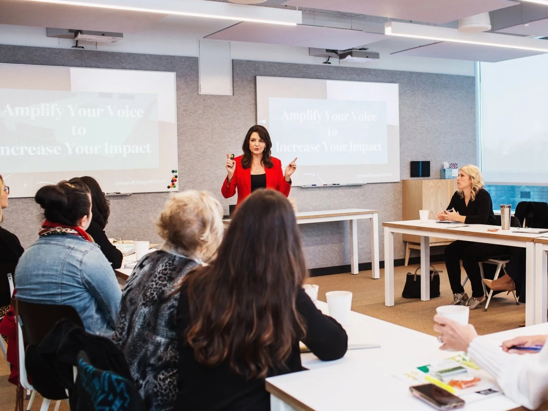 A woman in a red blazer is giving a presentation to a group of adults sitting at tables in a modern conference room. Two projection screens display the words 'Amplify Your Voice to Increase Your Impact.' The audience is attentively listening, some taking notes, with drinks and notebooks on the tables. The room is bright with a large window on one side.