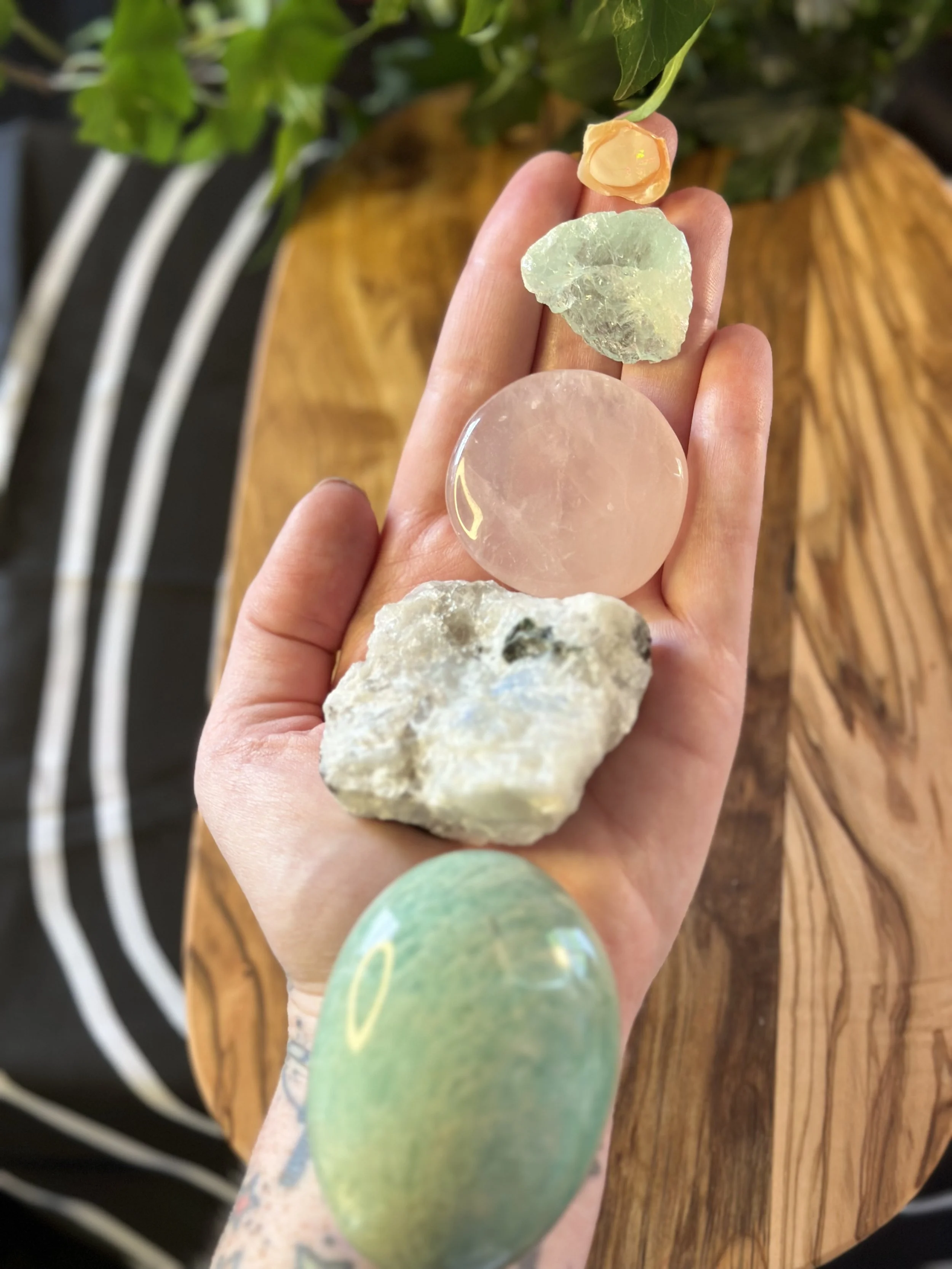 Person holding various stones and minerals, including aventurine, moonstone, rose quartz, aquamarine, and sunstone, with wooden and striped furniture in the background.