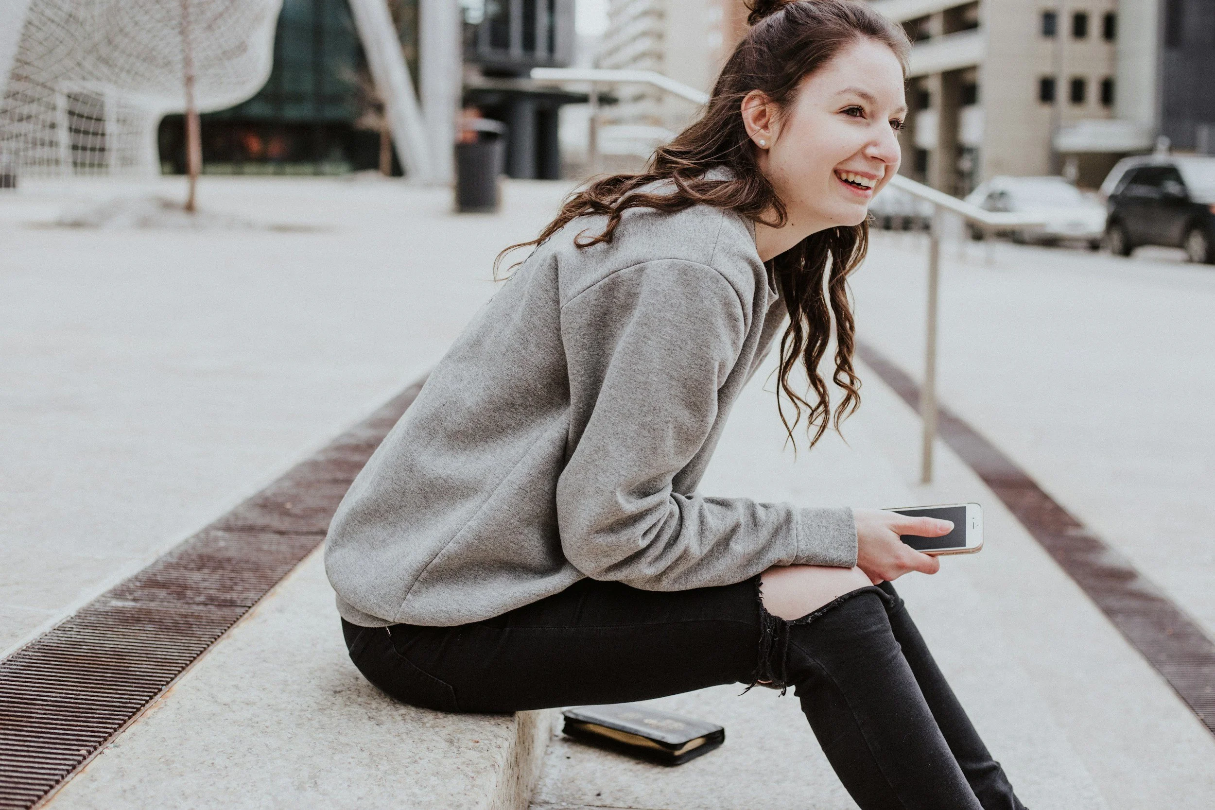 A young woman with long curly brown hair sitting on a park bench, holding a smartphone in her hand, smiling, wearing a gray hoodie and black pants, in an urban setting with buildings and parked cars in the background.
