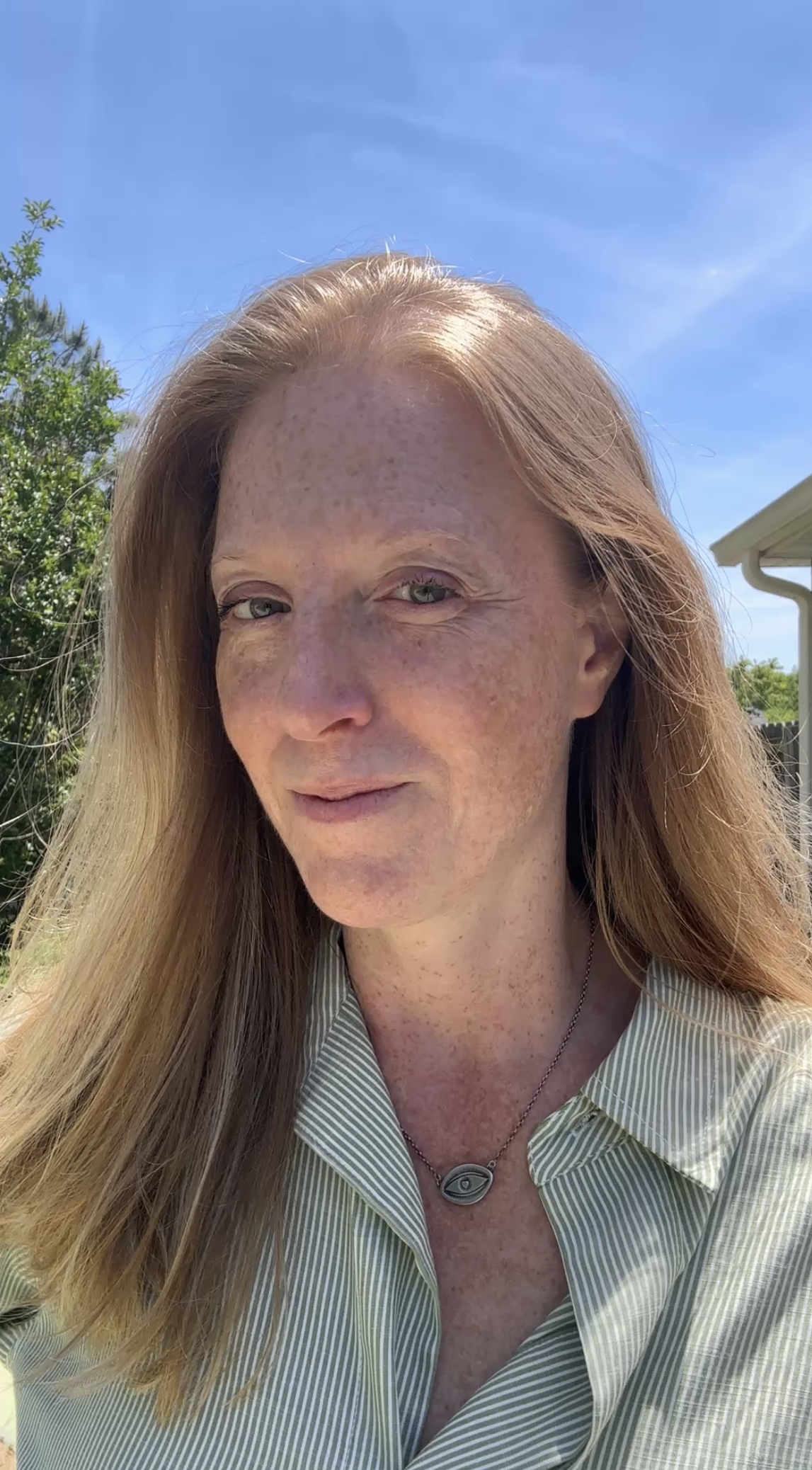 Close-up of a smiling woman with long red hair, wearing a striped shirt, a necklace with an eye pendant, standing outdoors under a blue sky with some greenery in the background.