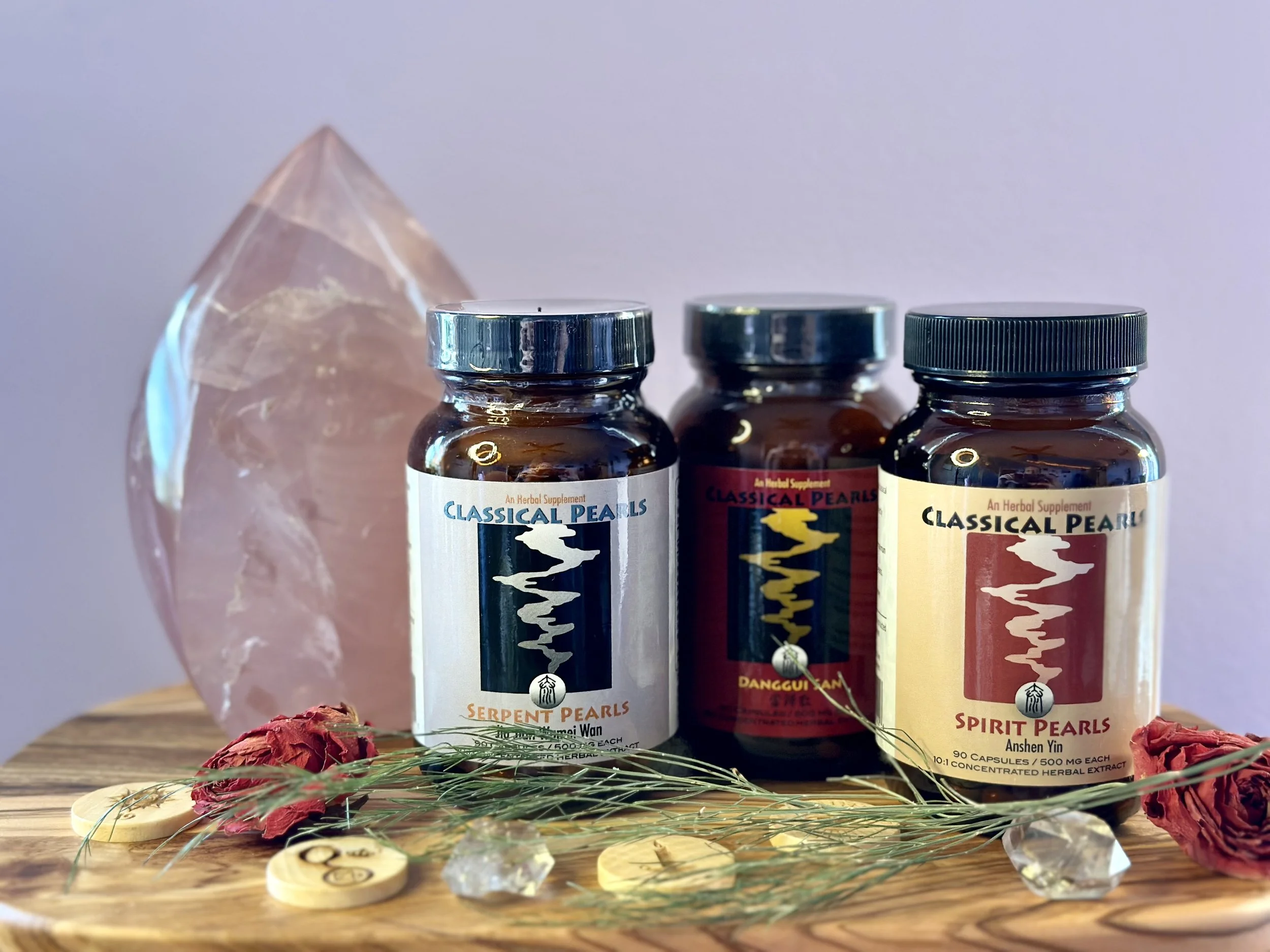 Three bottles of herbal supplements labeled 'Classical Pearls' in different varieties are placed on a wooden surface, with dried rose petals, slices of dried fruit, and herbs scattered around them. A large pink quartz crystal is behind the bottles.