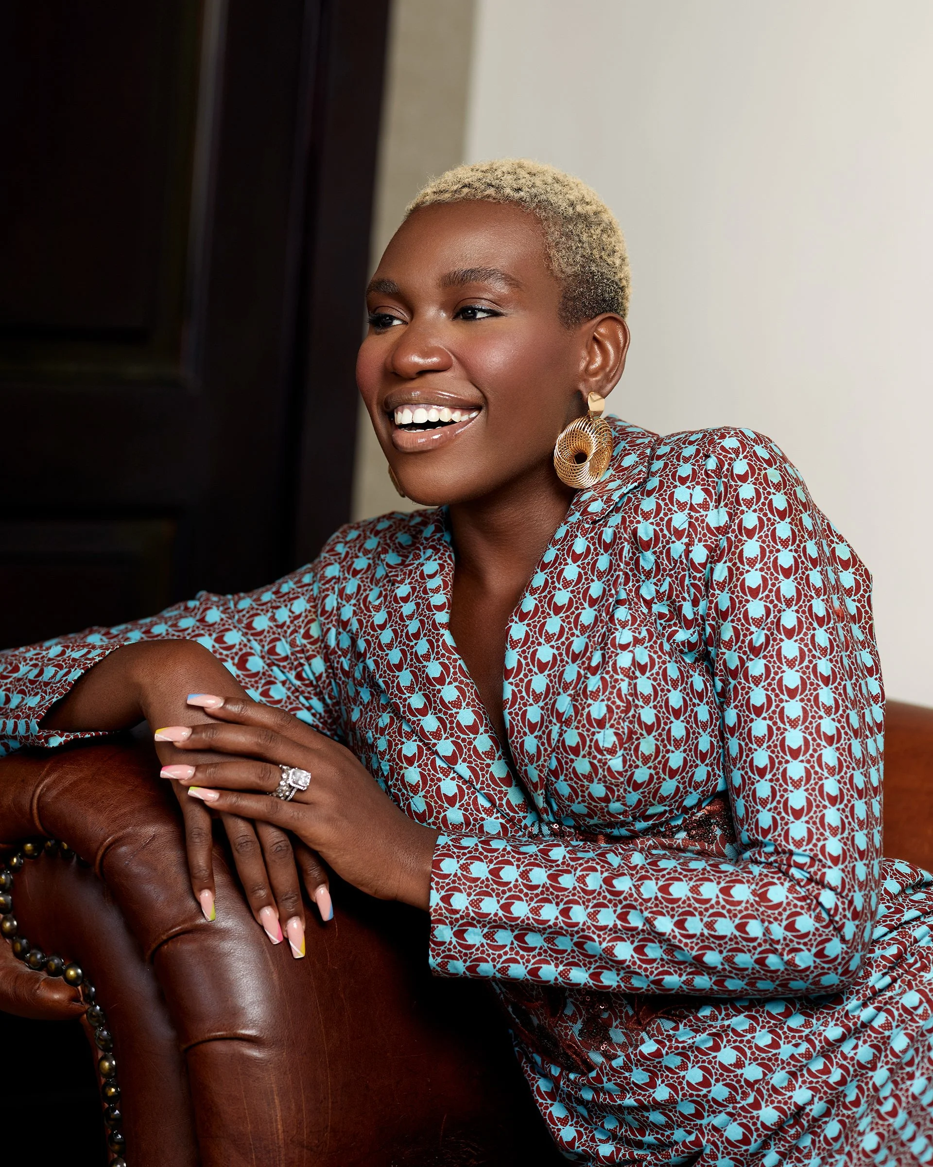 A woman with short blonde hair smiling, wearing a patterned dress with puffed sleeves, large earrings, and a wedding ring, sitting on a leather chair.