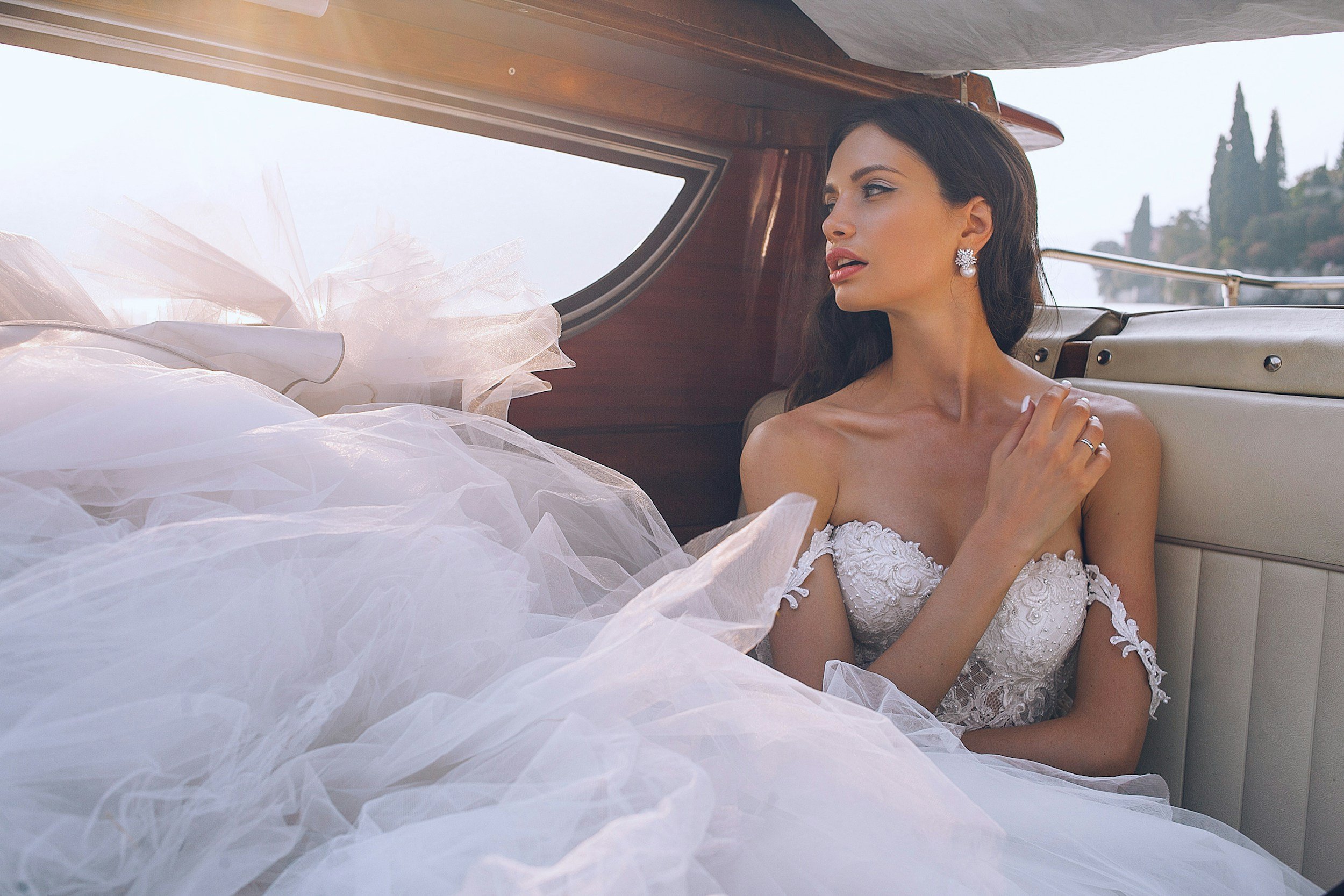 A woman in a white wedding dress with lace details sitting in the back of a classic car, looking out the window with a serene expression. The interior of the car has wood paneling, and the scenery outside features trees and a bright sky.