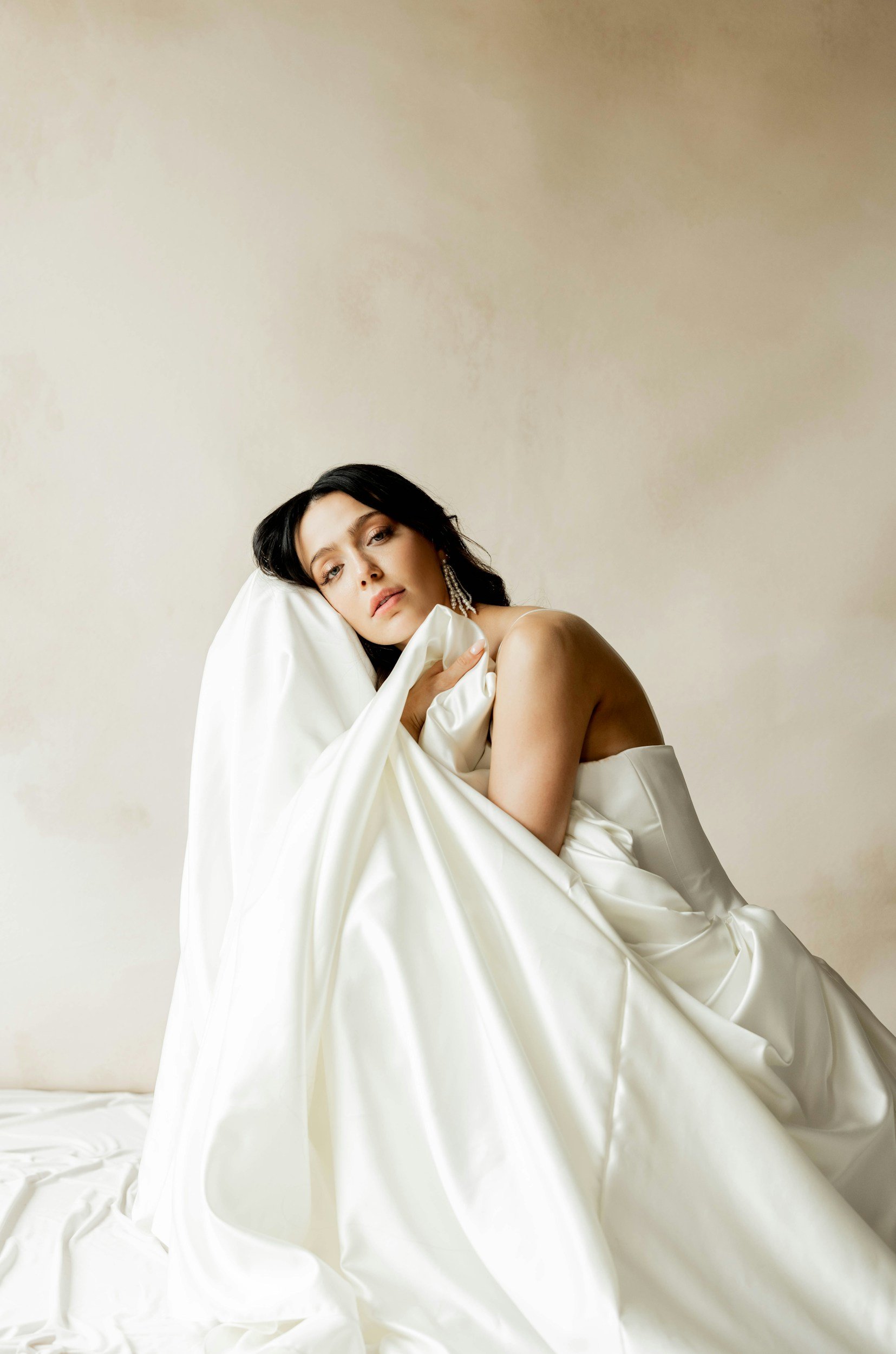 A woman with dark hair and earrings, partially covered with white satin fabric, posing against a plain beige background.