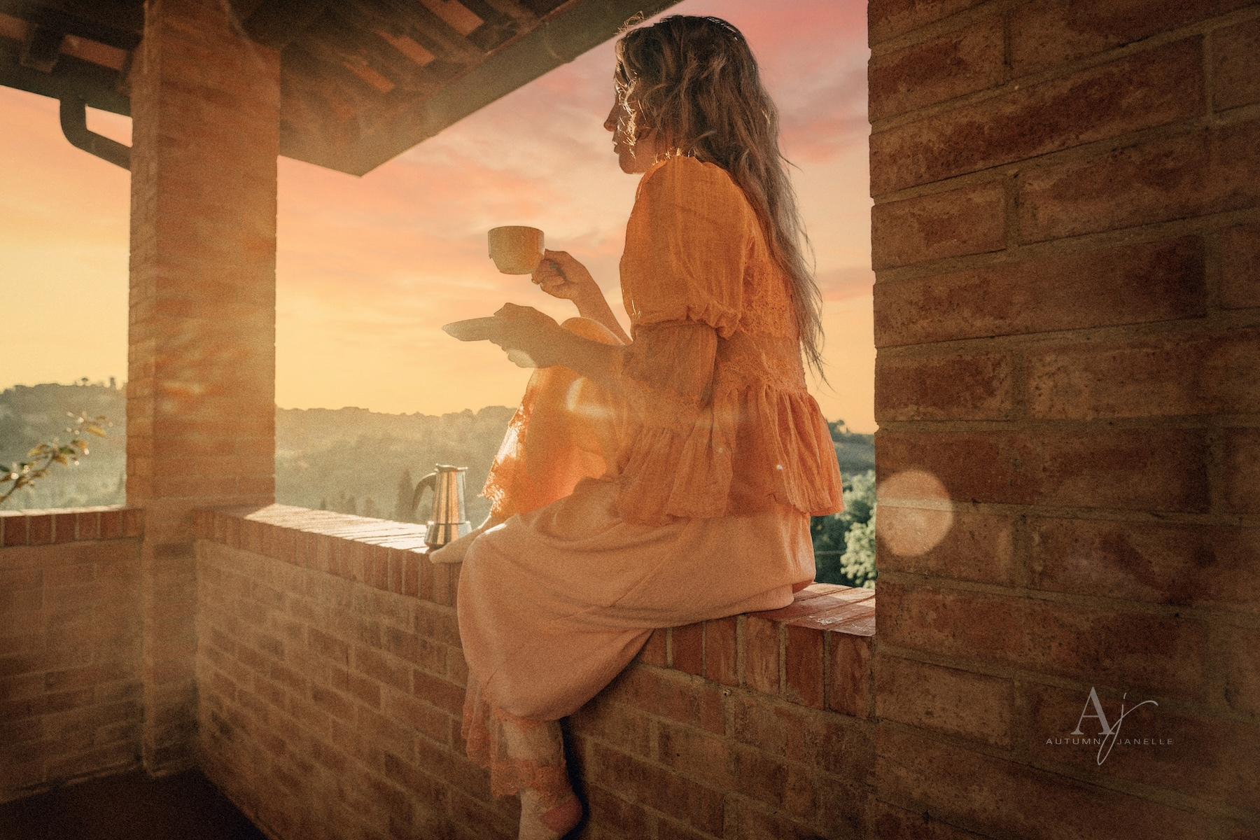 woman in lace top and bloomers sitting on ledge of Villa drinking coffee with San Gimignano in distance at sunrise