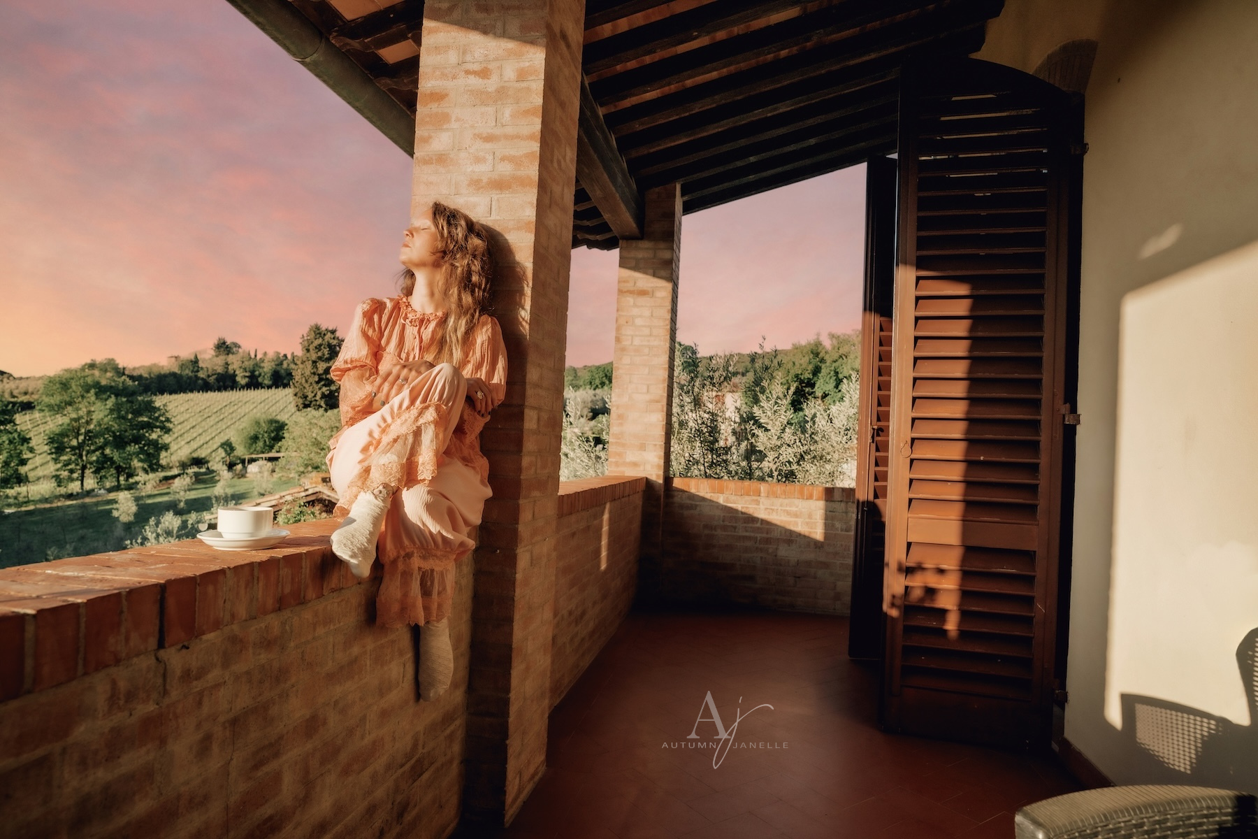 woman in lace top and bloomers sitting on ledge of Villa Raccienello drinking coffee with San Gimignano in distance at sunrise