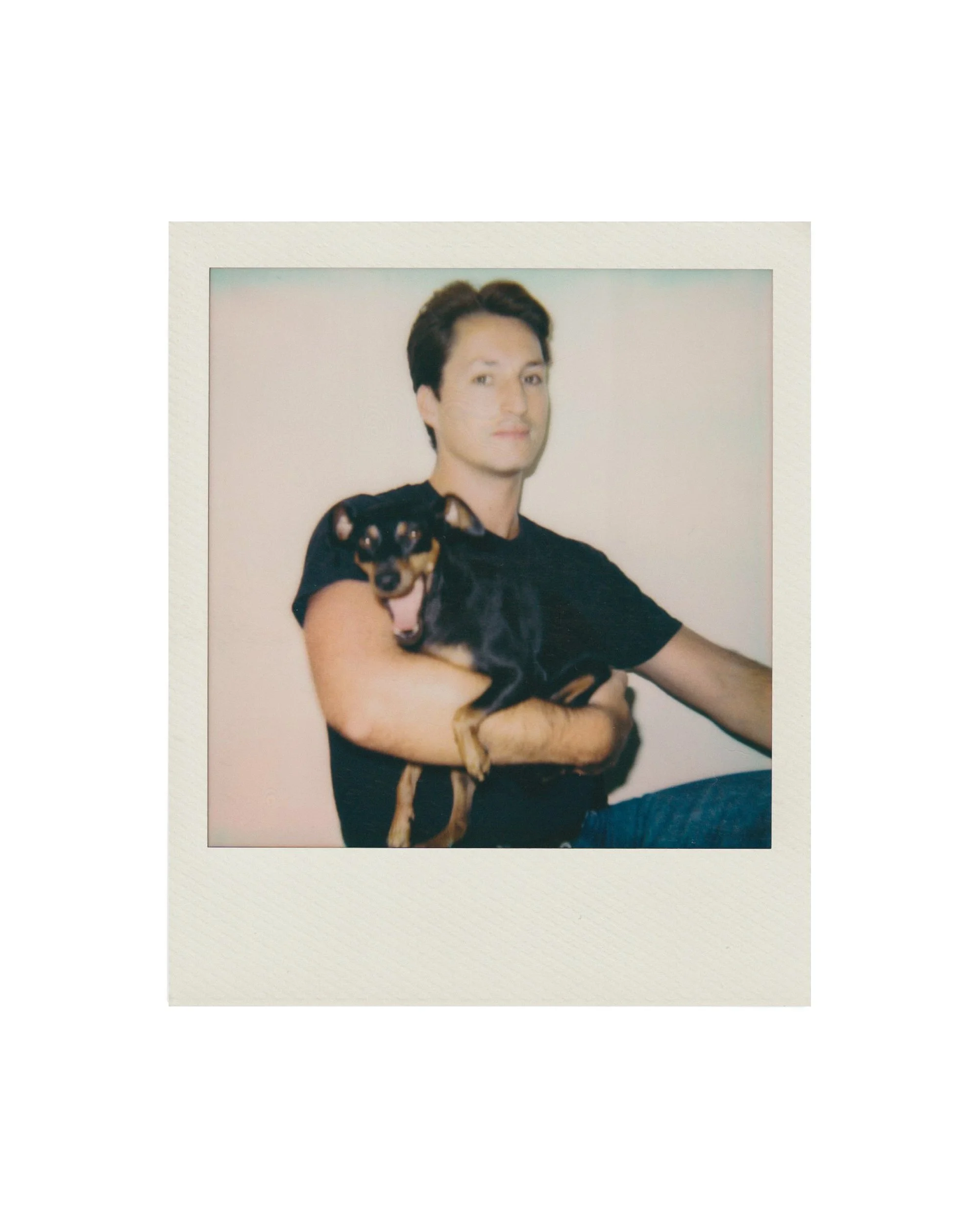 A man holding a small black and tan dog, both posing for a photo. The man is wearing a black t-shirt and has short dark hair.