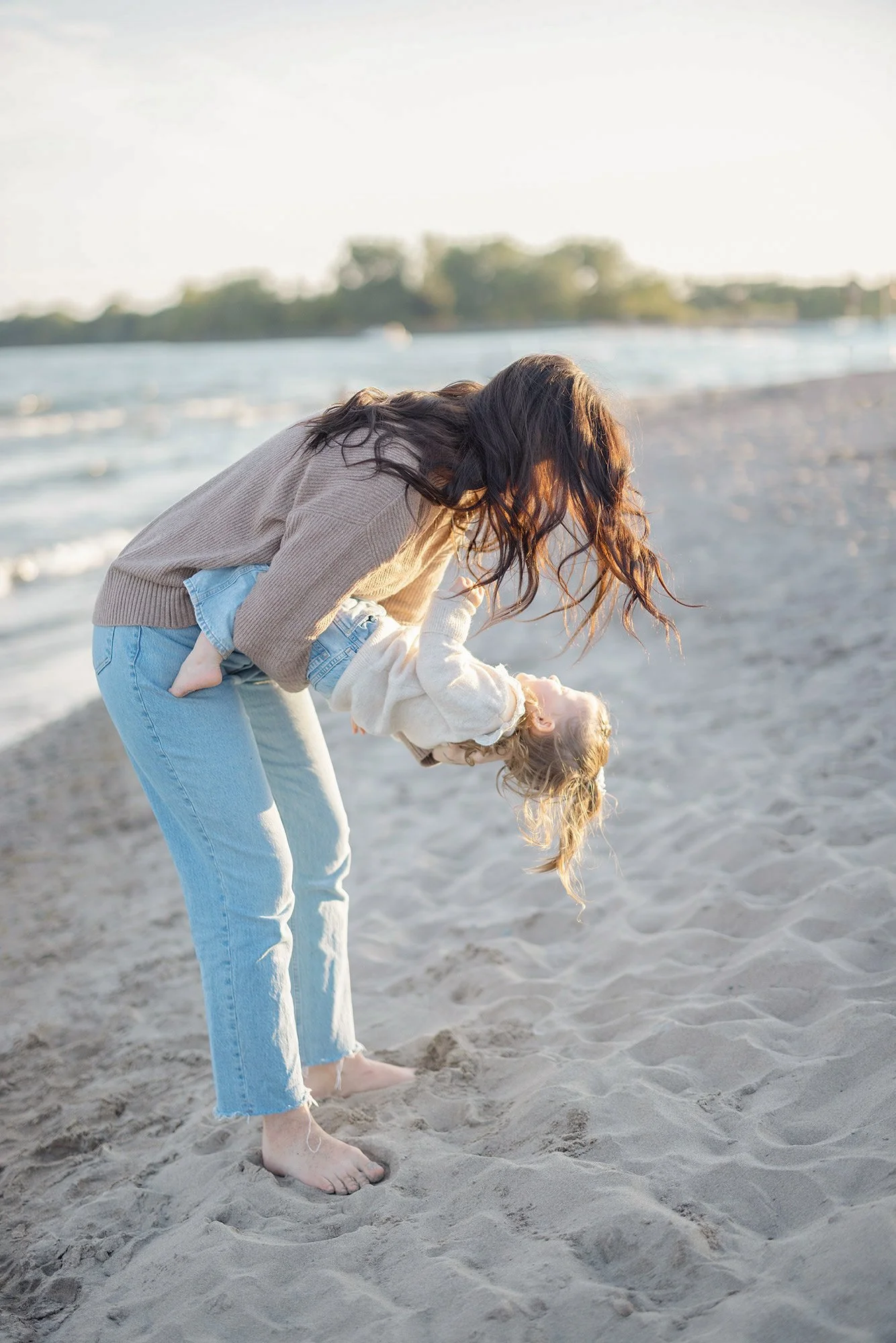 candid photo of woman holding child on beach. Toronto skyline in the distance. 
