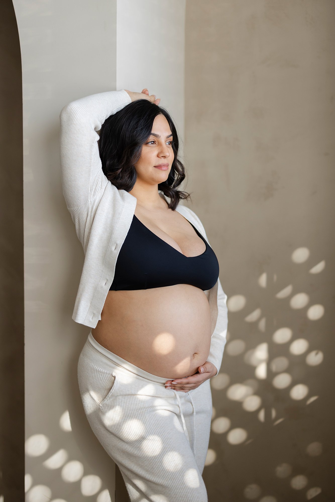 in studio maternity photo. Mom is wearing neutral clothing and leaning against the wall.