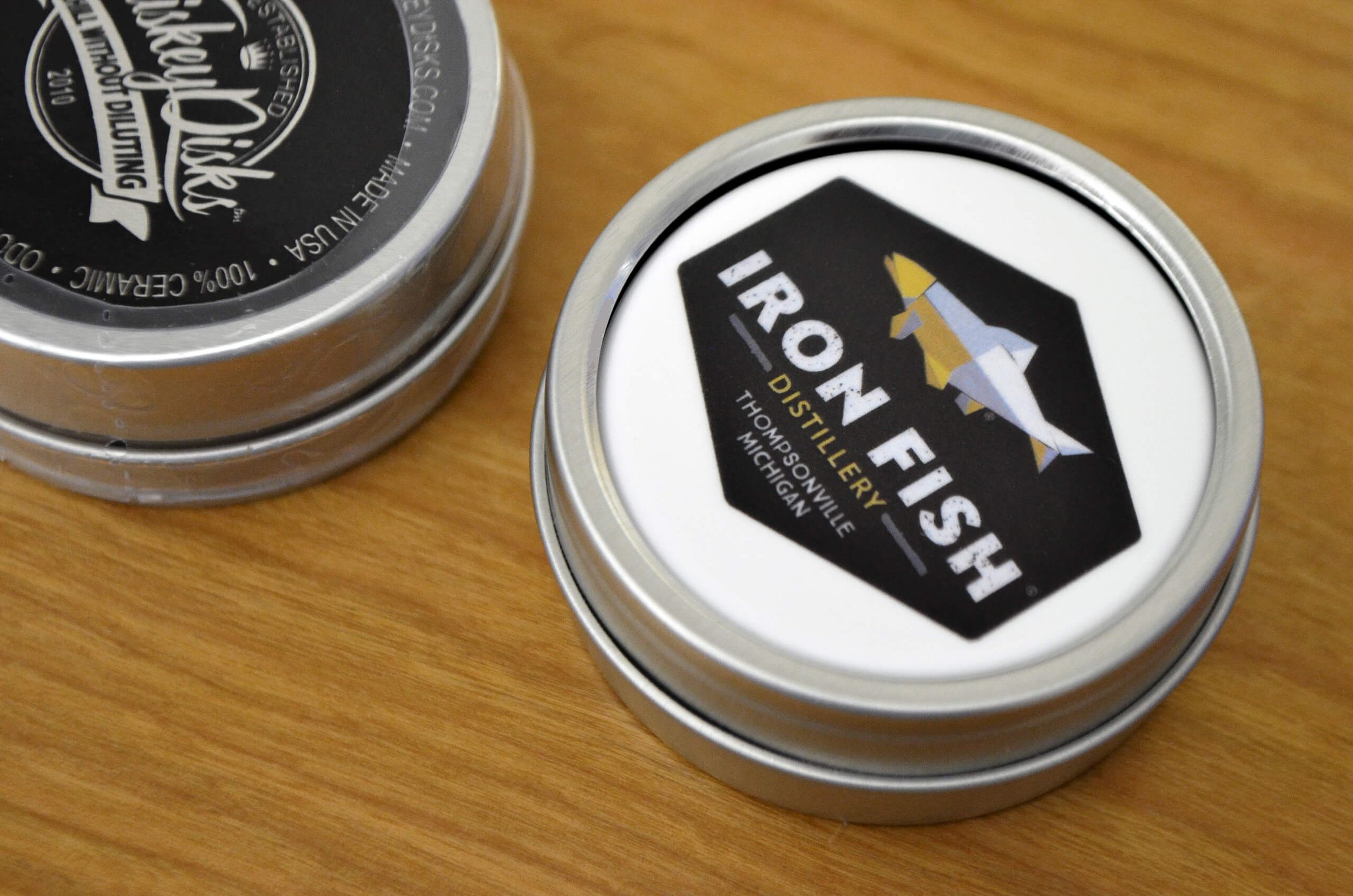 Iron Fish branded Whiskey Disk packaged in silver tin sitting on a wooden tabletop. Additional unside down packaged Whiskey Disk to the side, showing product information on the bottom of the tin.