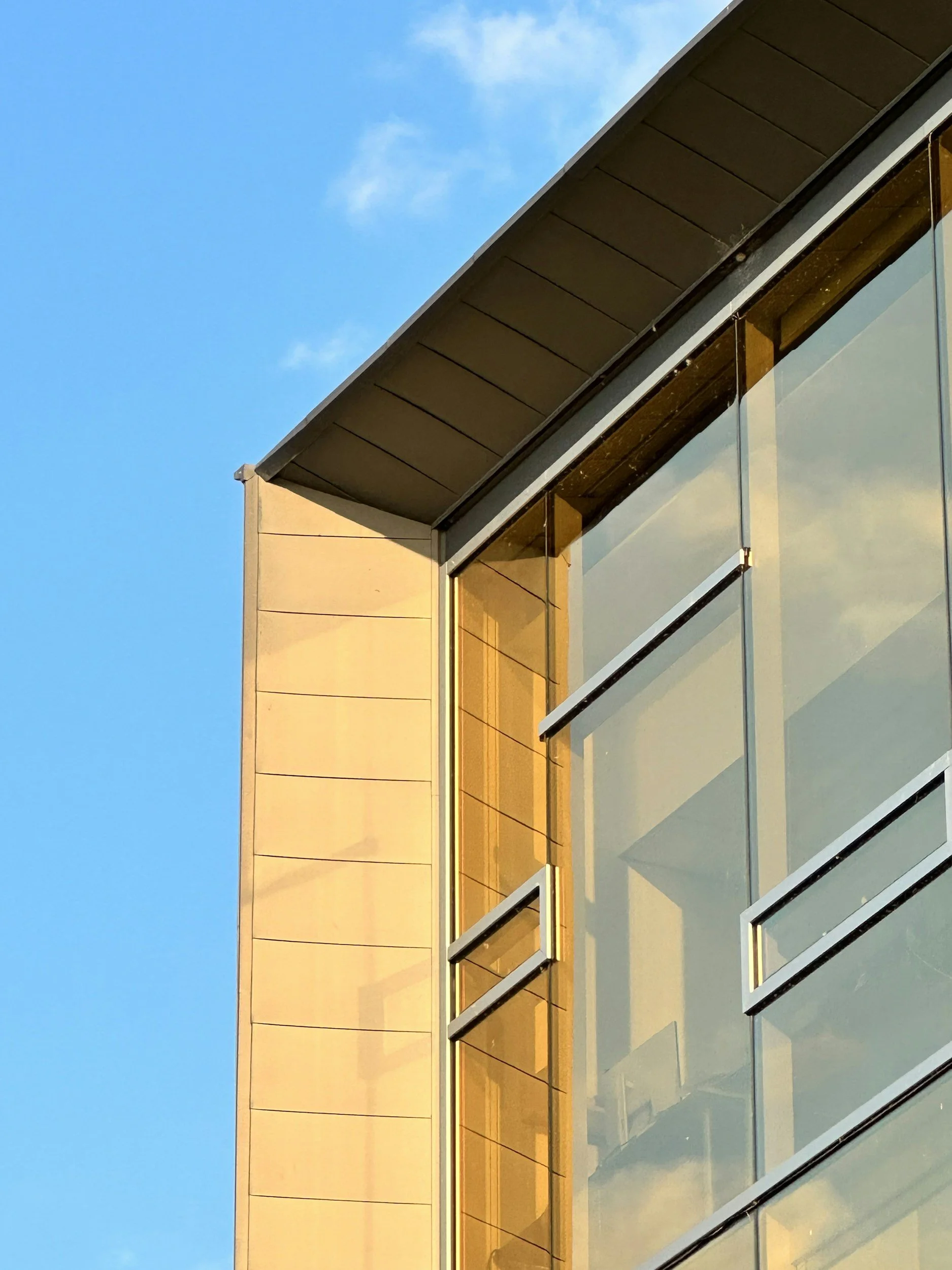 Close-up of a modern building corner with glass windows, beige panels, and a blue sky background.