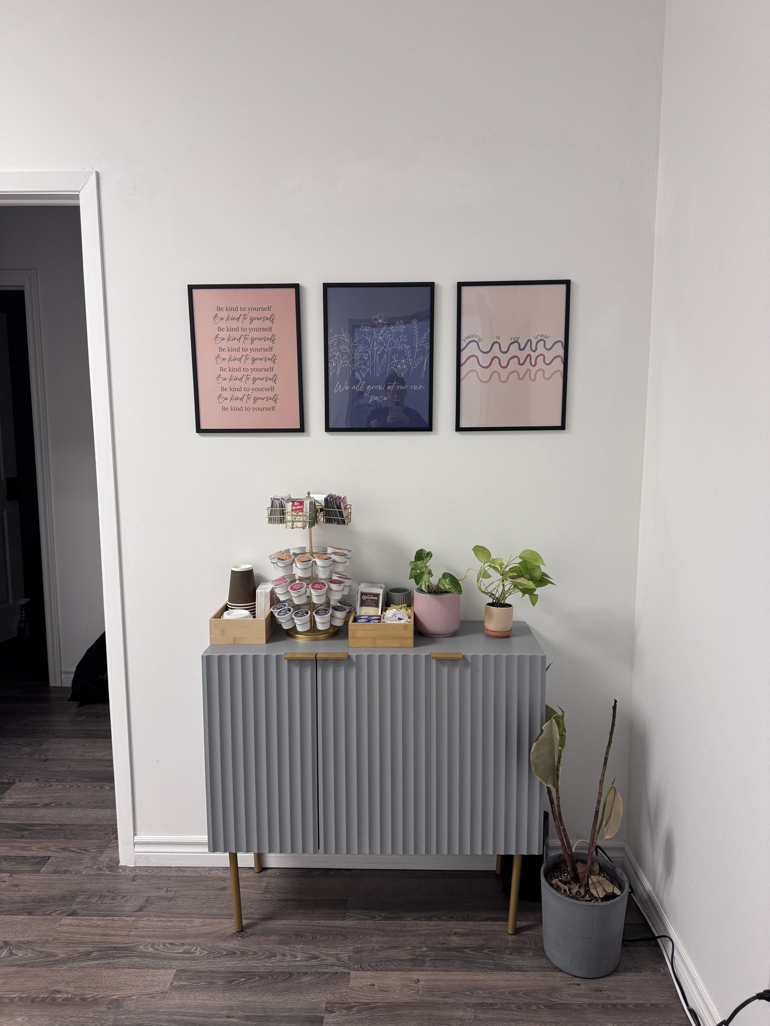 A gray sideboard with coffee pods and tea supplies on top, two potted plants, and three pink, blue, and black framed art prints on a white wall above.