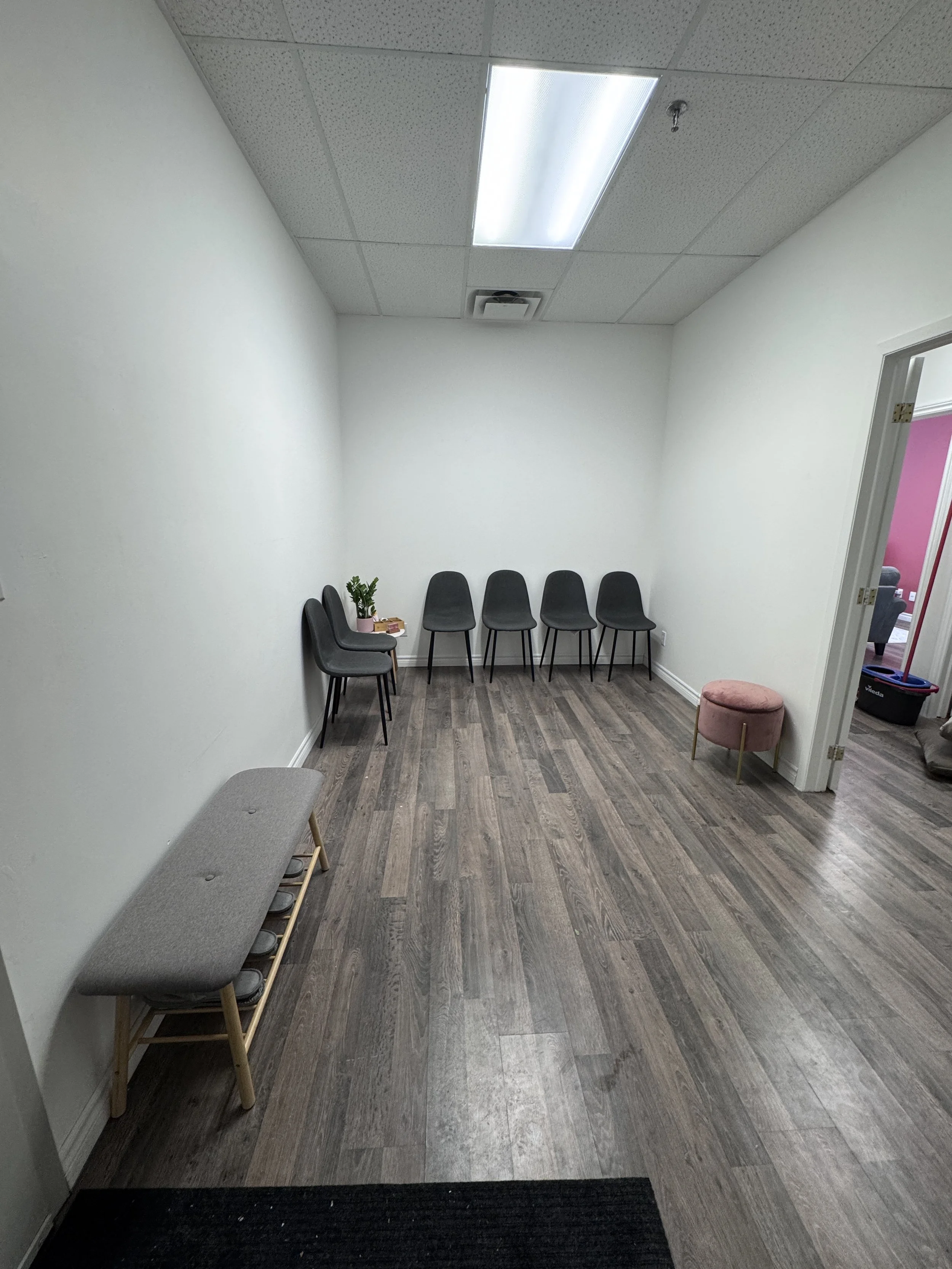 Empty waiting room with five black chairs, a pink plant, a grey bench, a pink ottoman, and an open door showing another room with a red wall.
