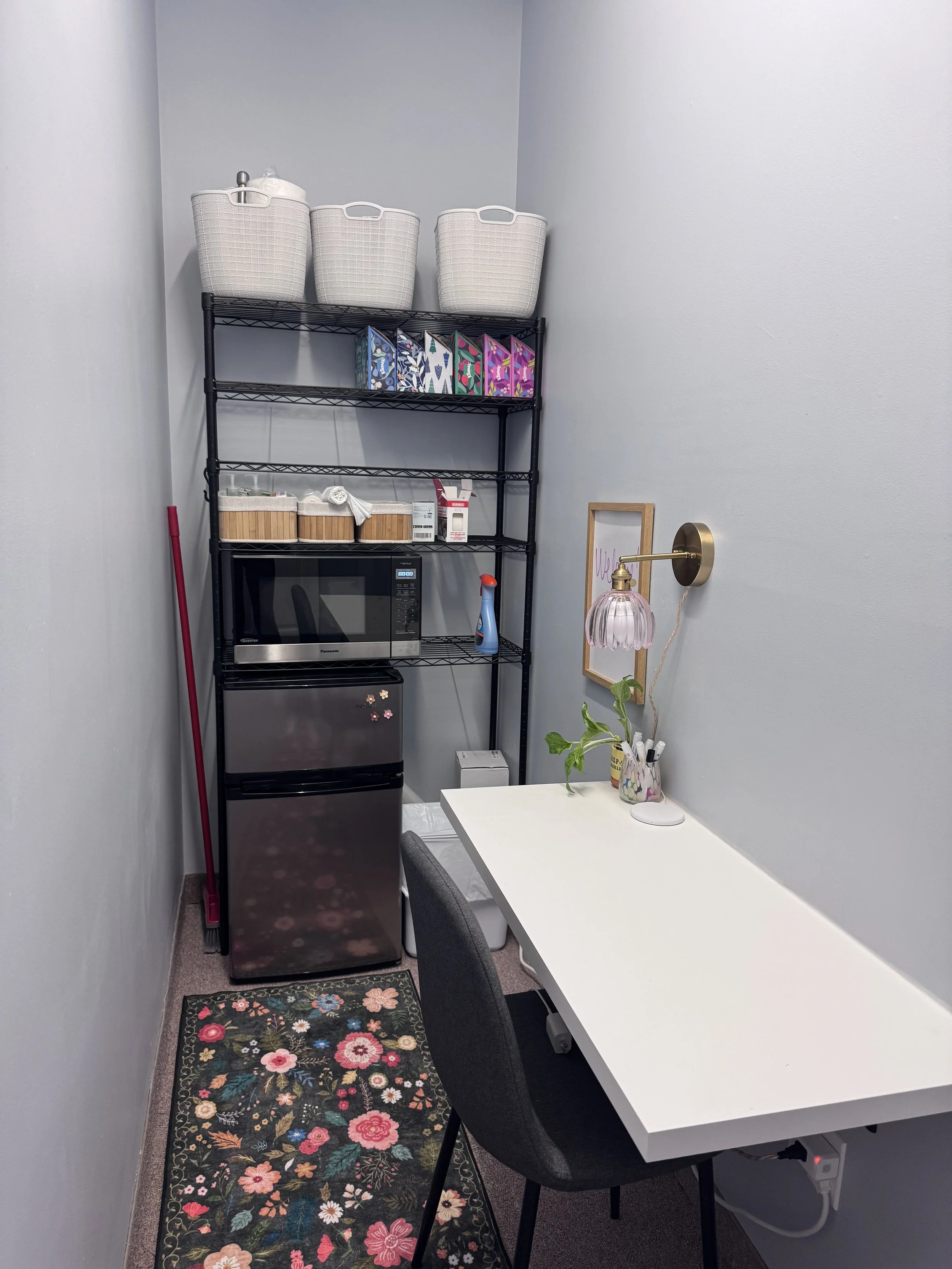 A small room with a shelving unit, microwave, mini fridge, and report on a white table with a pink lamp and a plant.