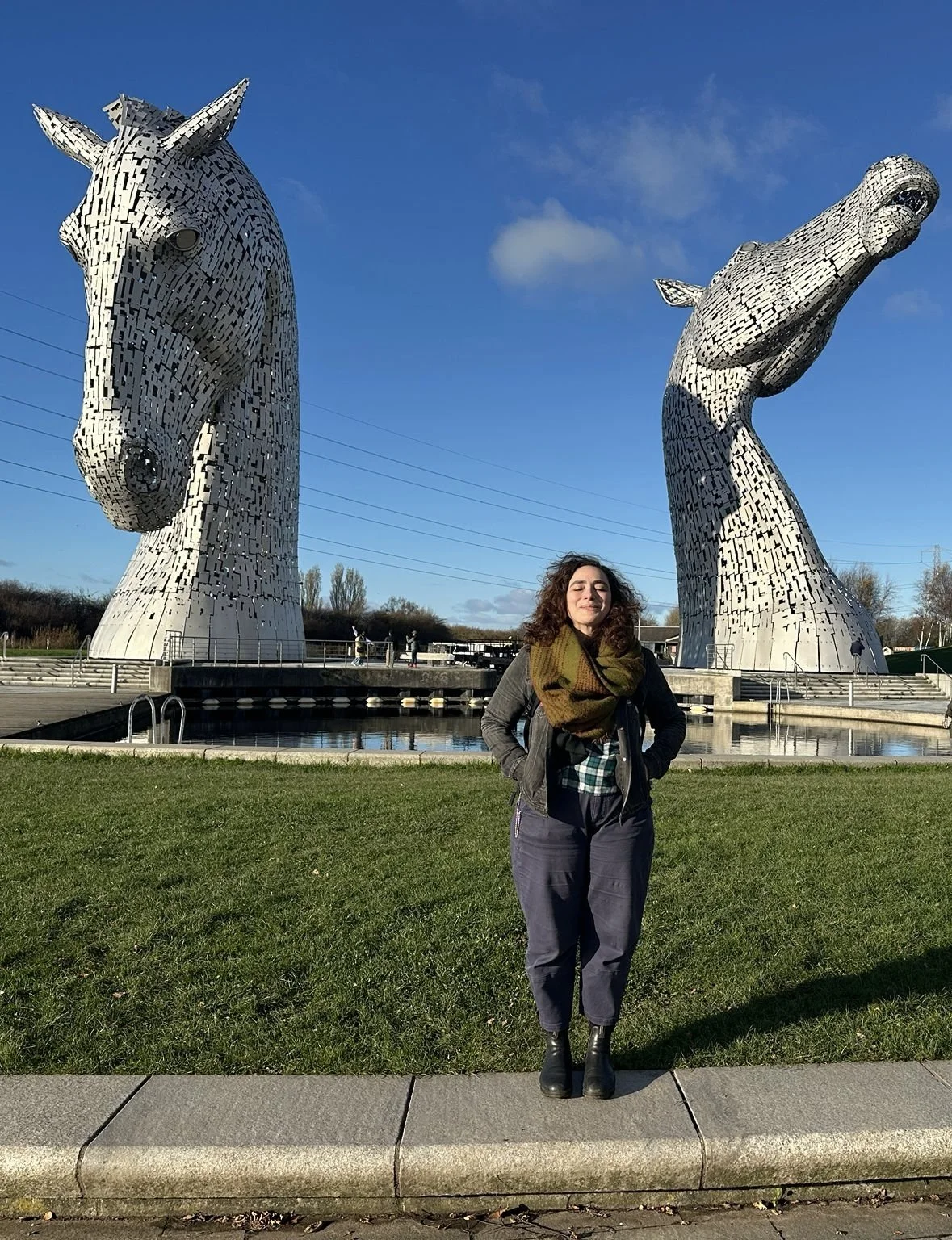 Catherine Blake Smith stands in a field, in front of public artworks of two massive sculptures of two horse heads.