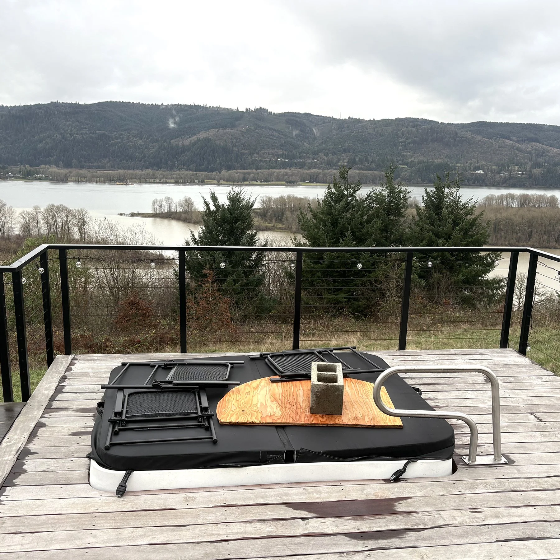 Photo of a hot tub installed on a deck with a cover on it, and a cinder block and piece of plywood on top to keep the cover from blowing off