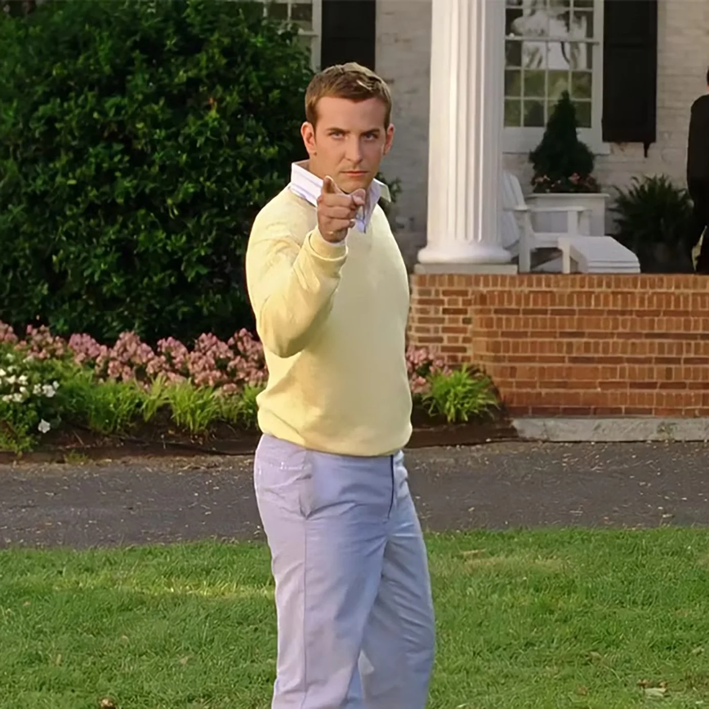 Still shot of Bradley Cooper from Wedding Crashers. He's wearing a yellow preppy sweater and blueish pants standing outside a very nice house,  pointing at the camera