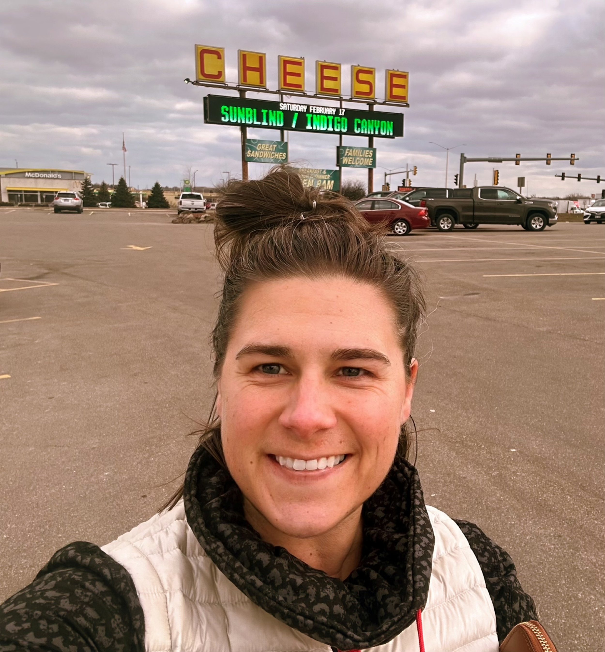 Jamie Carpenter stands in a parking lot in the foreground with a sign in yellow, with red lettering, reads "Cheese" in the background.