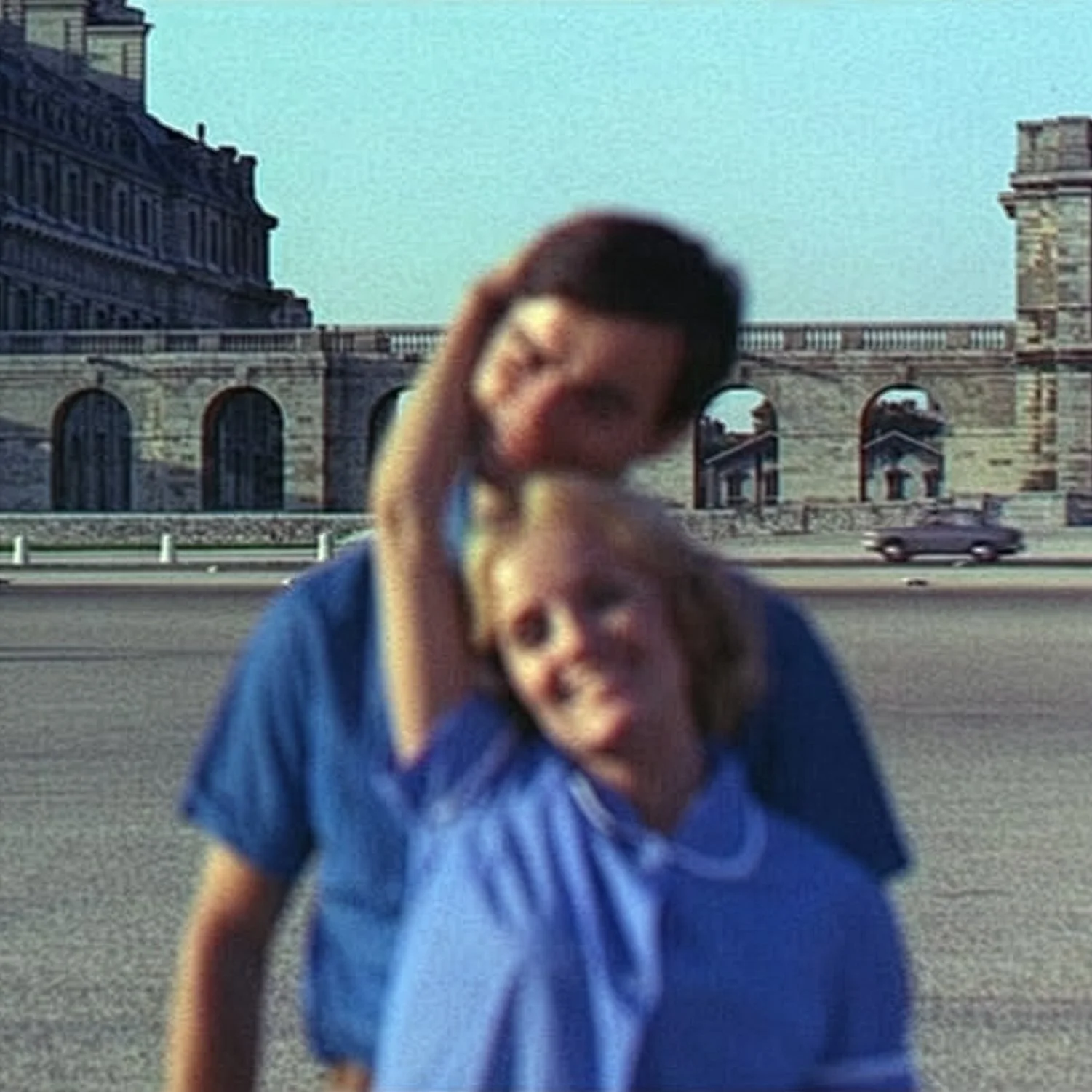 Image from the French film Le Bonheur that shows a male/female couple, both wearing blue shirts, posing outside in Paris. The building in the background is in focus, but the main characters are blurry, as if the camera focused  on the wrong thing.