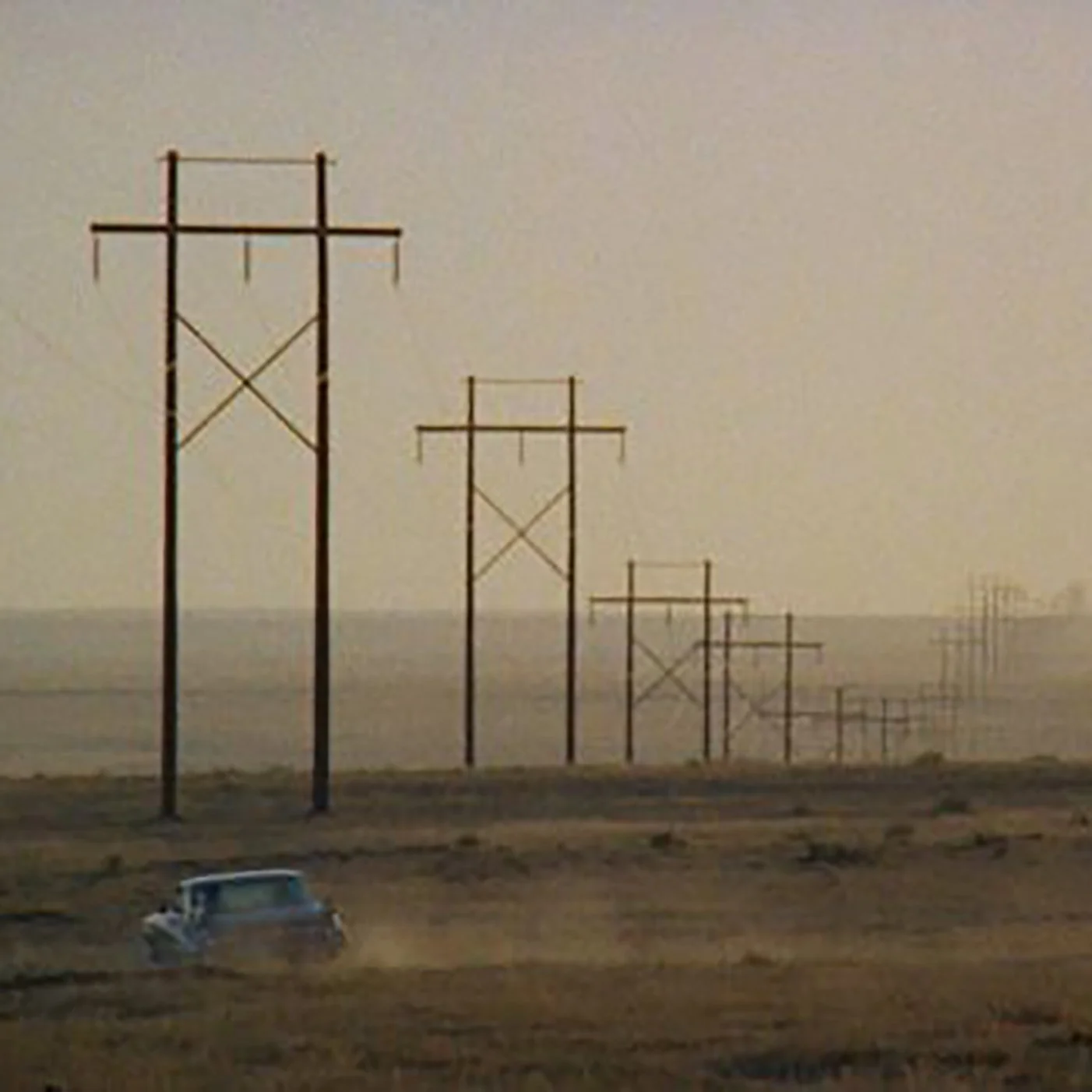Screen shot of the movie the bandlands that is a very wide angle, zoomed out image of miles and miles of dusty, dirty road with nothing around except power lines and a car driving in the distance kicking up dust