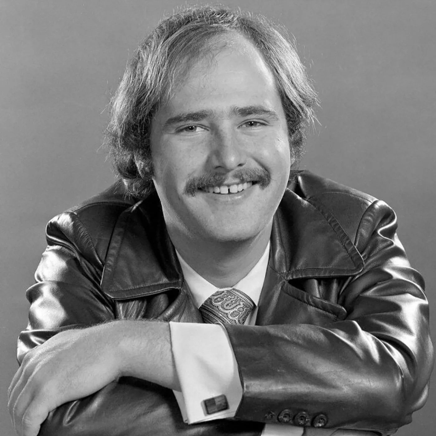 Black and white heaadshot  of Rob Reiner. He's wearing a leather jacket, sitting backwards in a chair, his arms cross in front of him on the chair back, and smiling. He has a mustache and shaggy-length hair
