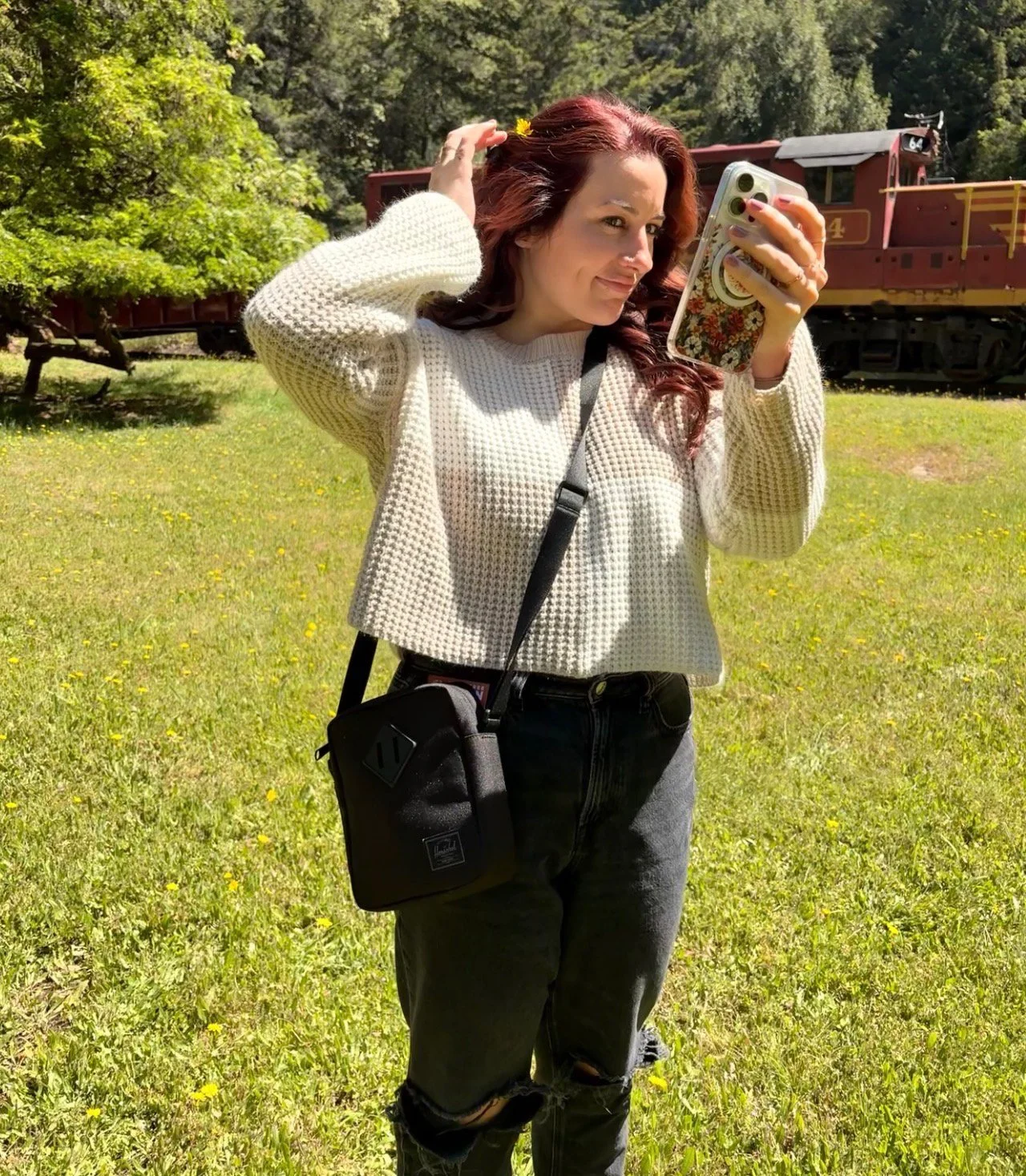 A photo of Shannon Moore standing in a field with a red train in the background, with her hand to her hair as she takes a selfie.