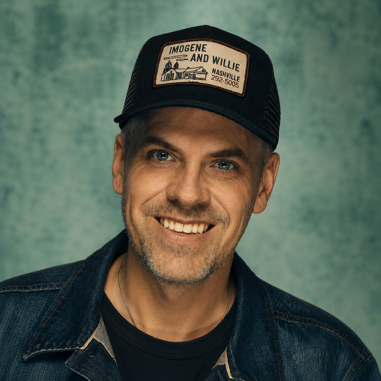 A headshot of Drü, who has a big smile and is wearing a hat that reads "Imogene and Willie. Nashville 292-5055."